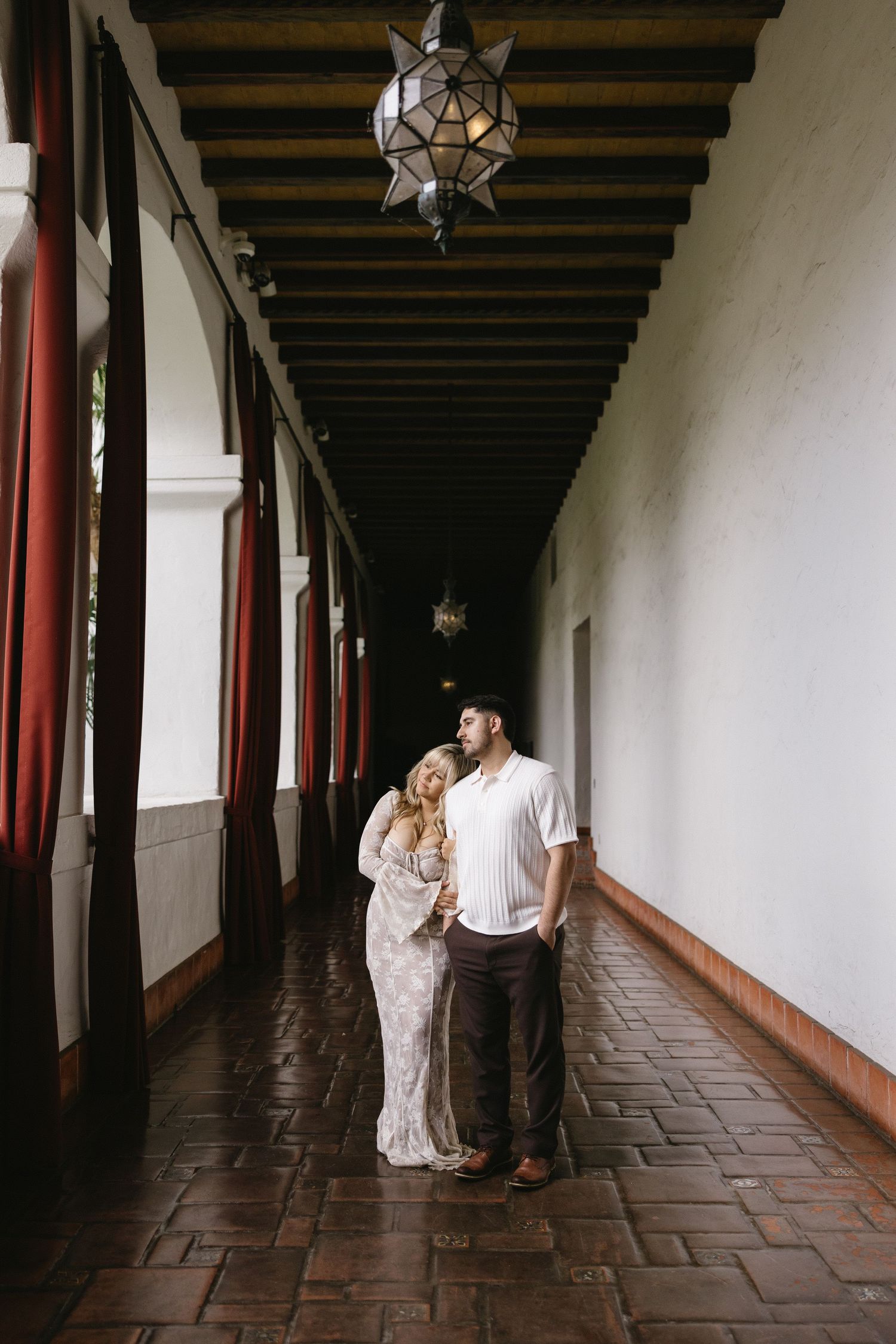 A couple walks through a Spanish colonial corridor with dark wooden beams and hanging star lanterns.