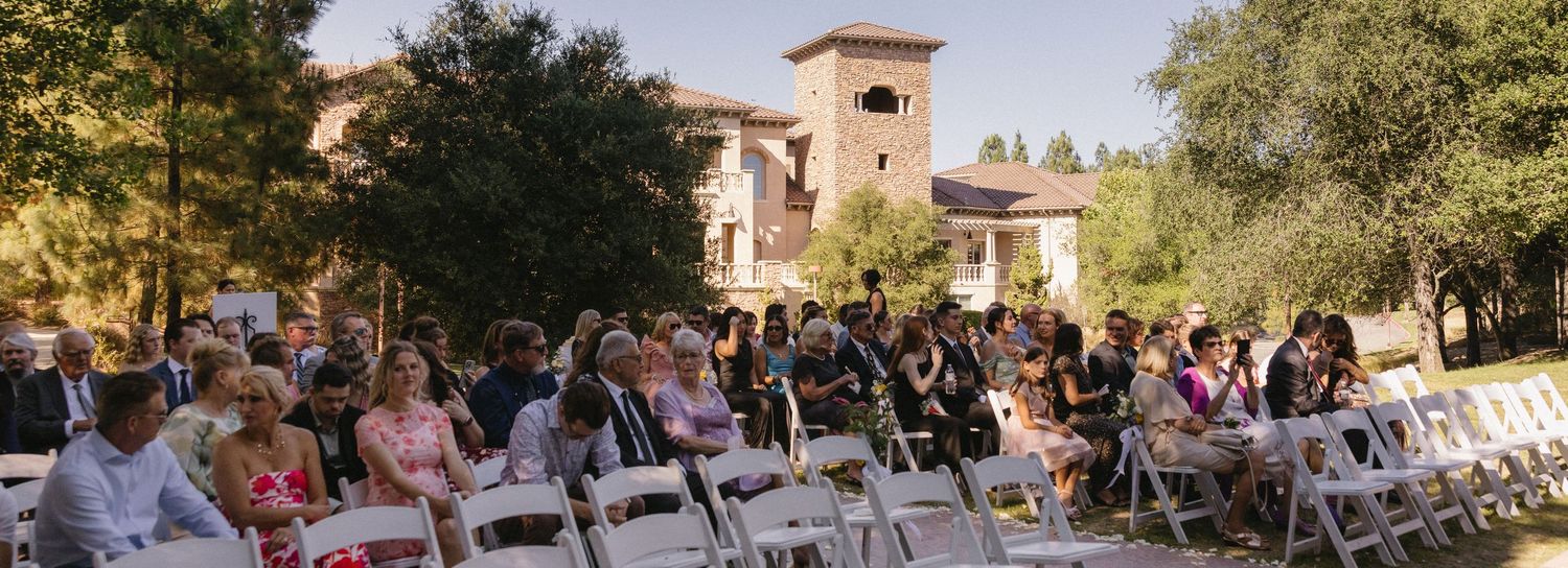 Outdoor wedding ceremony with guests seated on white chairs in front of a elegant stone villa surrounded by lush trees.