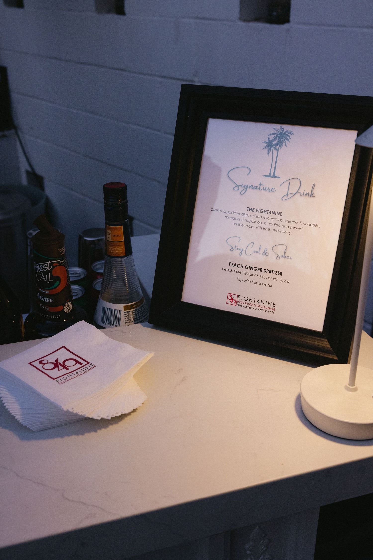 Framed signature drink menu on white bar with cocktail napkins and bottles at an elegant event.