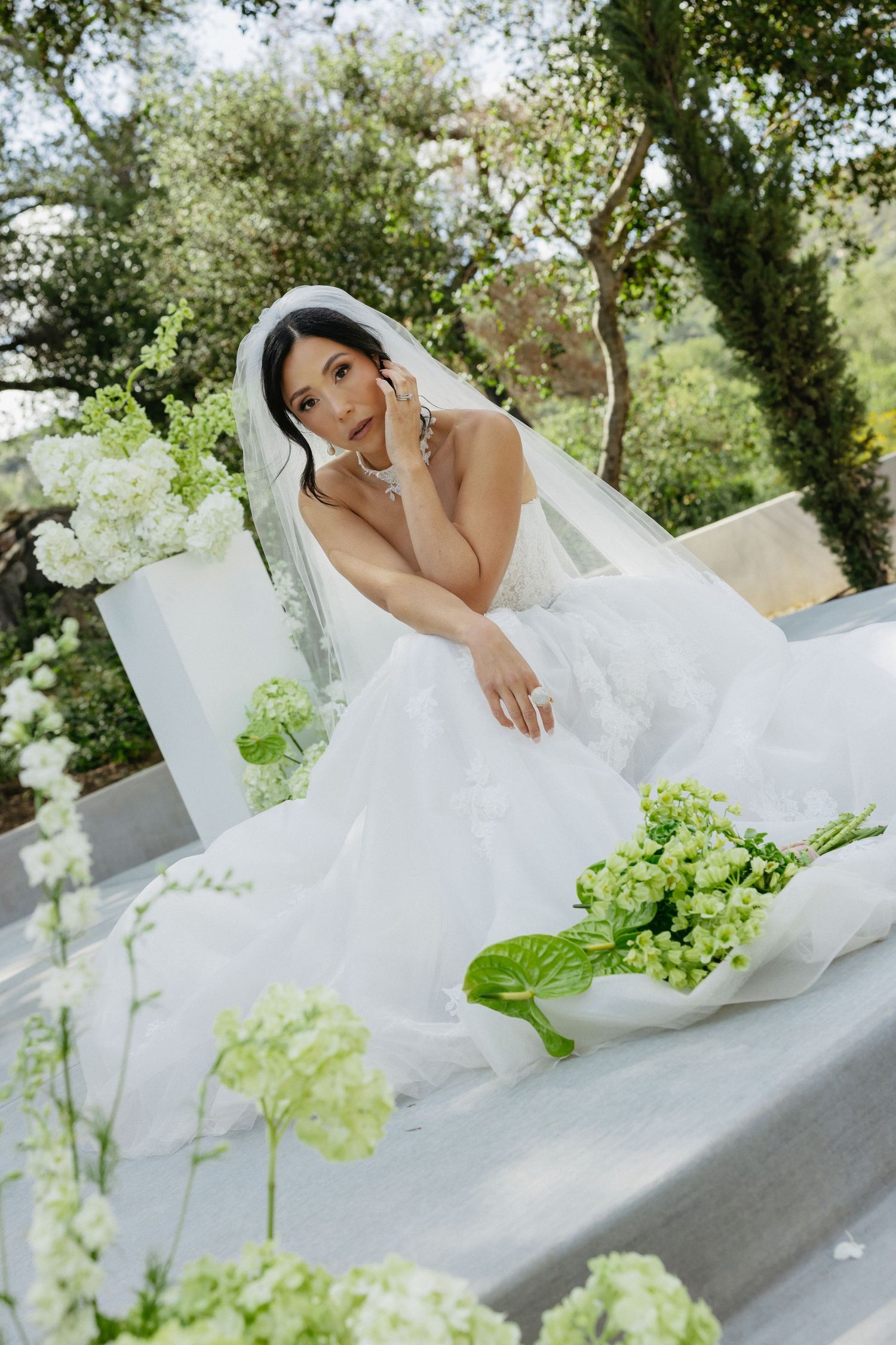 Elegant bride in white gown and veil seated among lush green and white floral arrangements outdoors.
