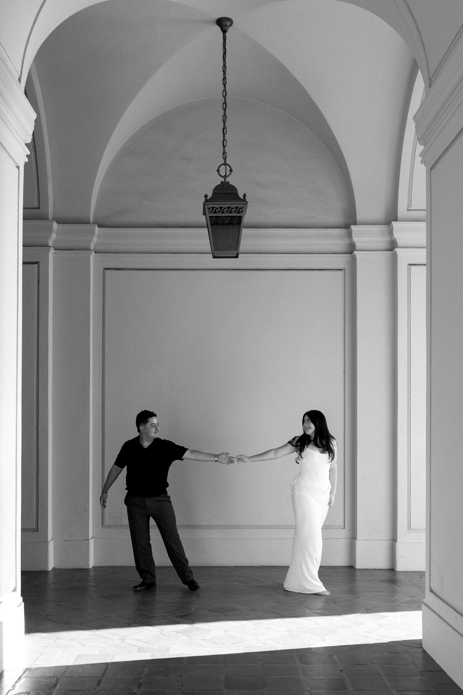 Black and white photo of a couple holding hands under an elegant arched hallway with a vintage lantern.