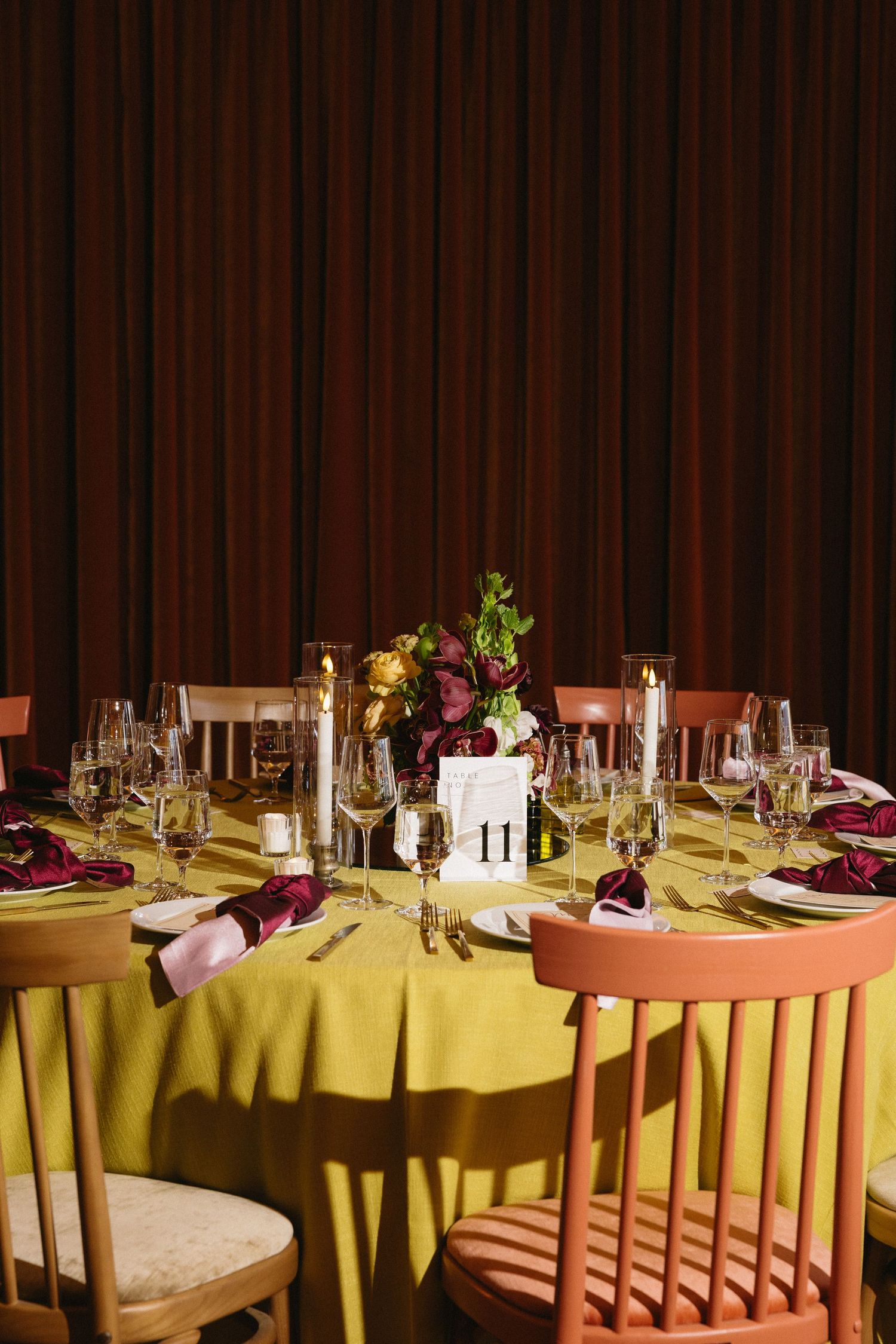 Elegant wedding reception table with yellow linen, burgundy floral centerpiece, candles, and mixed chairs against dark curtains.