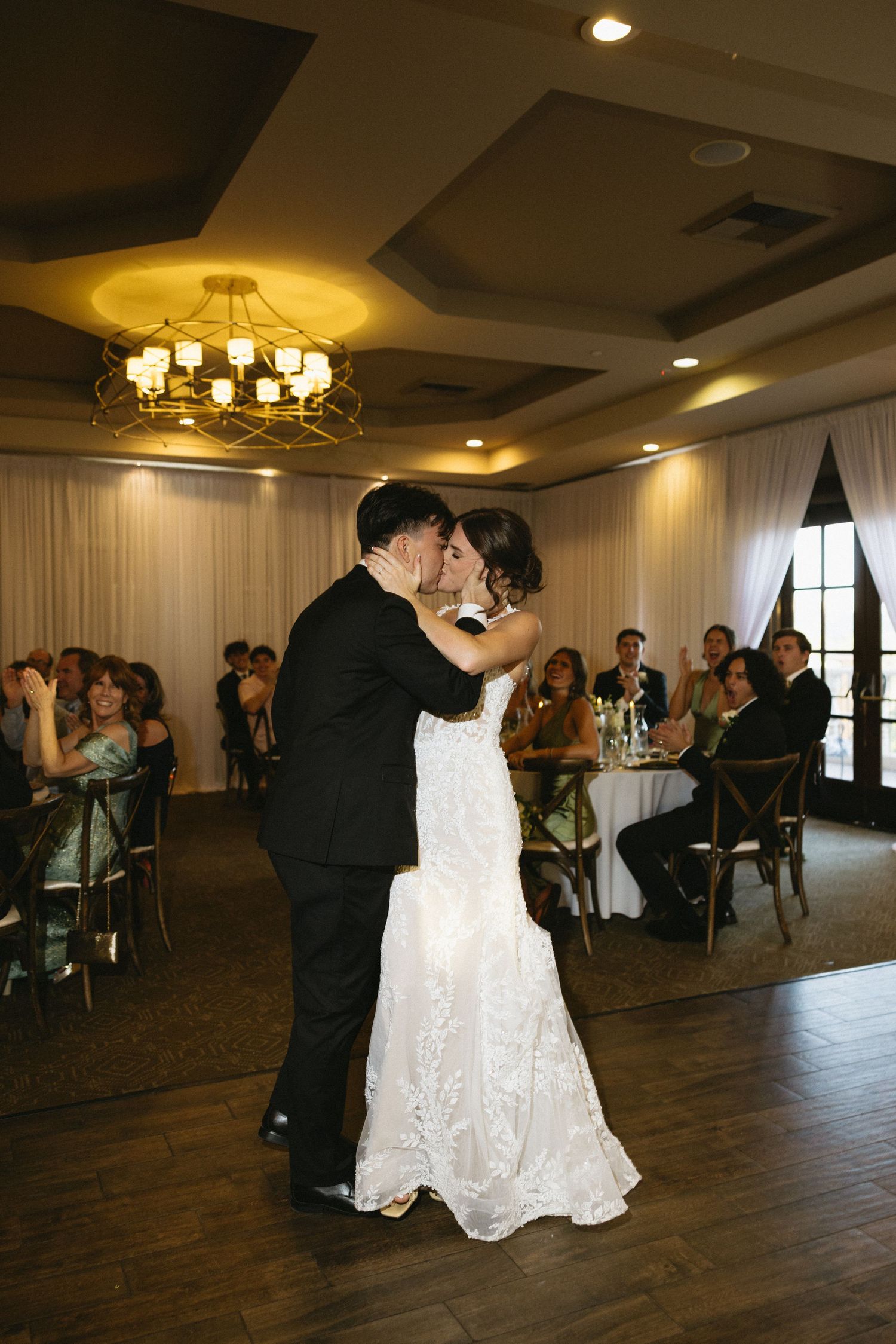 Bride and groom share their first dance at elegant wedding reception with chandelier and guests seated nearby.