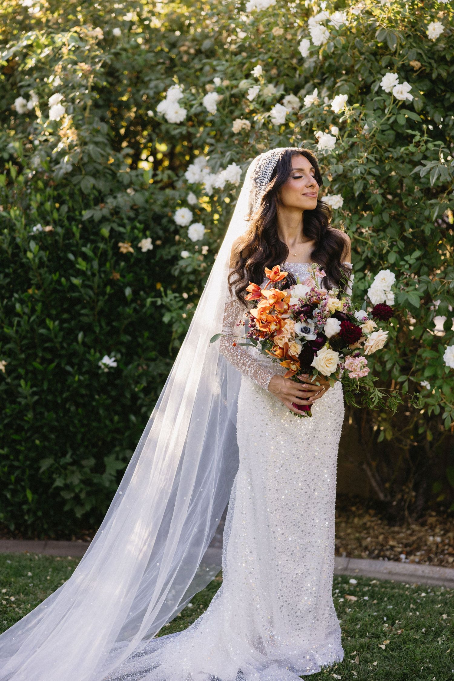 Bride in a lace gown with long veil poses in a garden with white and peach roses.