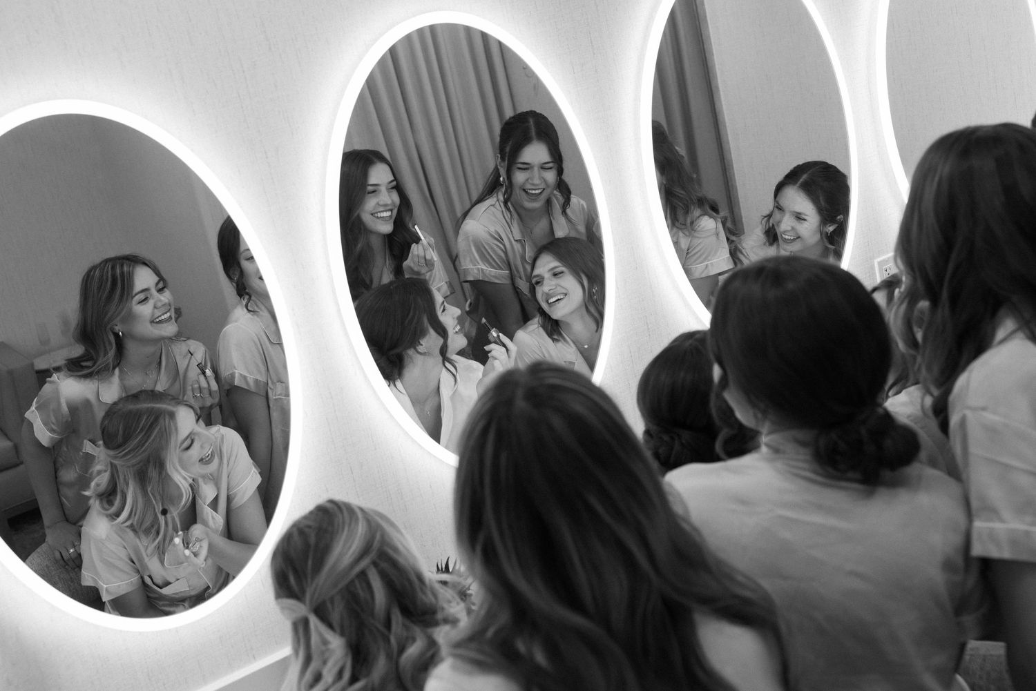 Bridesmaids laughing and getting ready together, reflected in oval backlit mirrors in black and white photo.