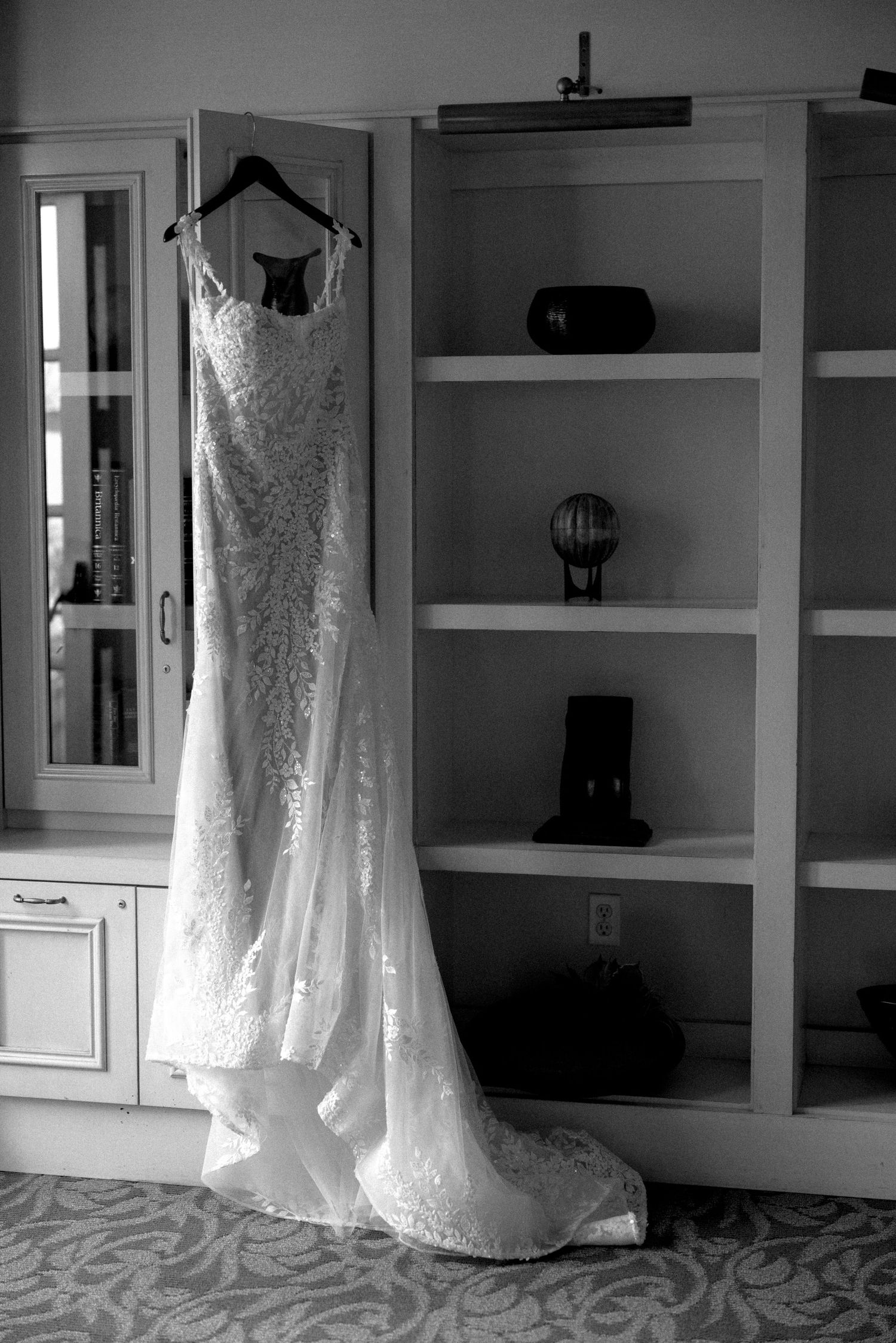 Black and white photo of an elegant lace wedding gown hanging on a hanger beside a decorative bookshelf.