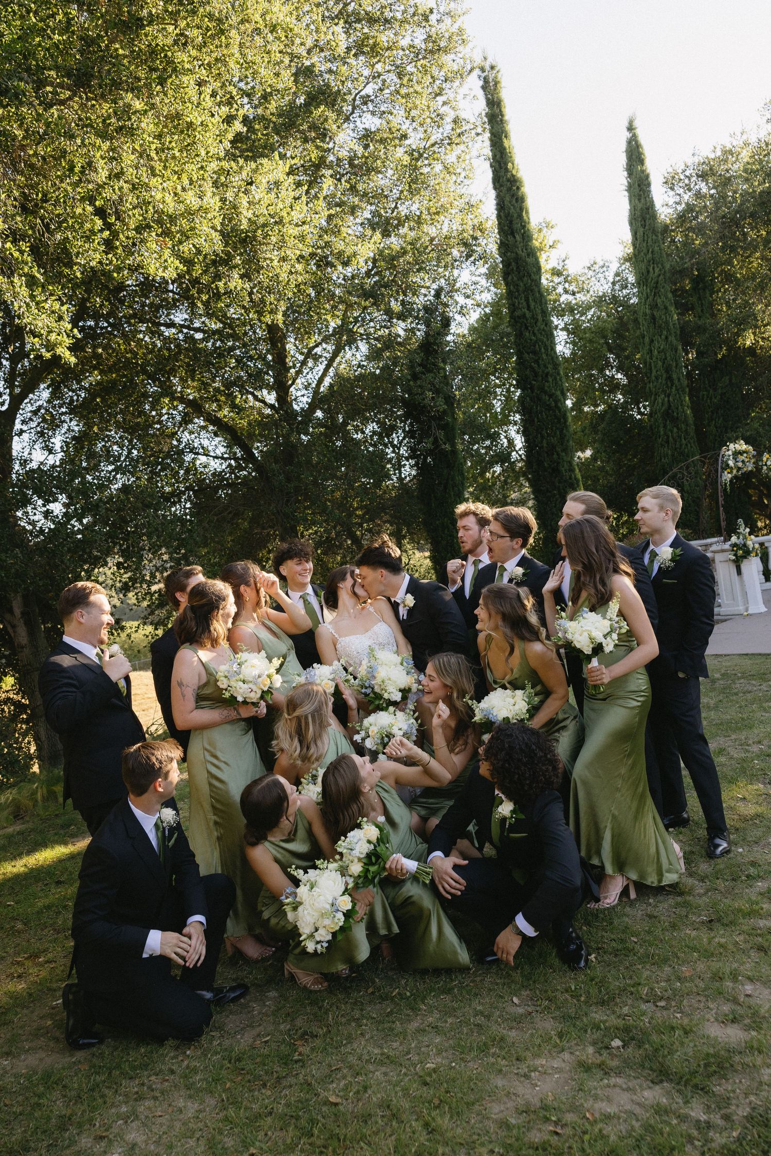 Wedding party in sage green dresses and black suits pose outdoors surrounded by cypress trees with white floral bouquets.