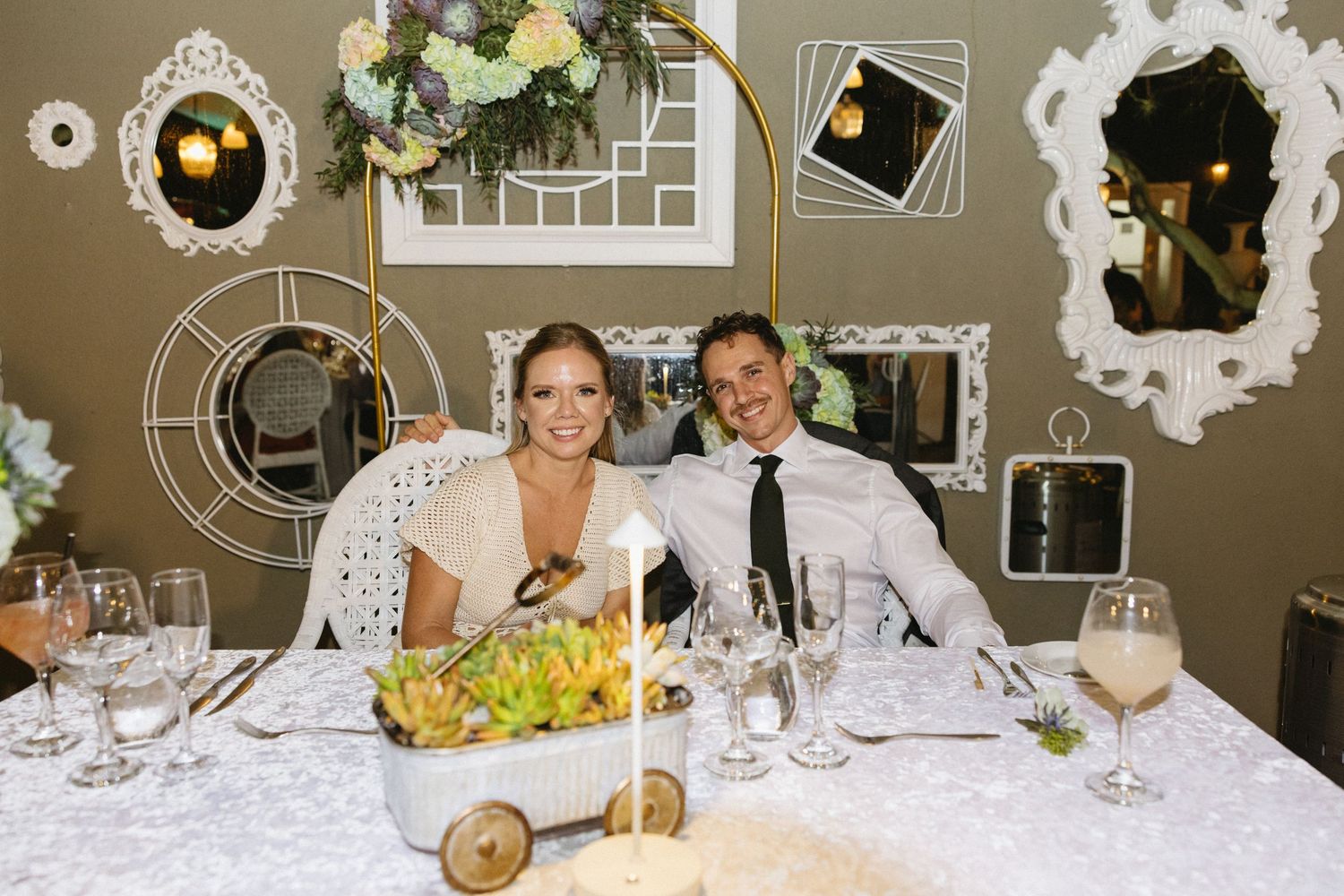 A smiling bride and groom sit together at an elegantly decorated wedding reception table with white lace and floral centerpieces.