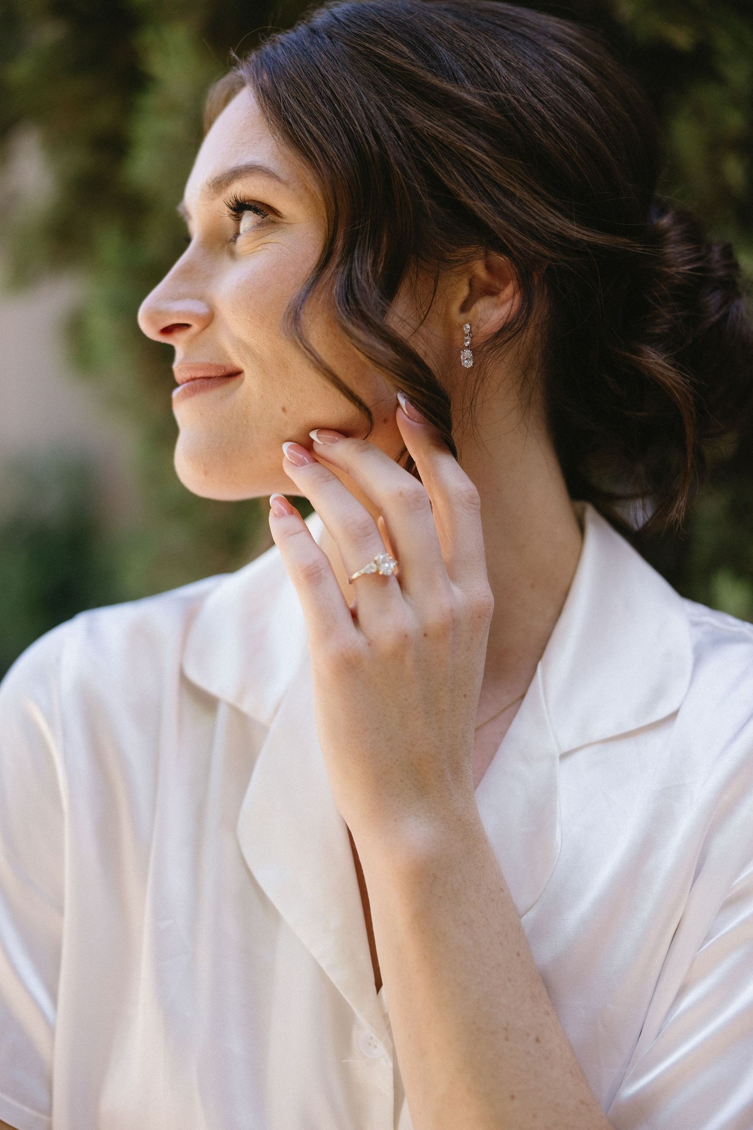 A smiling brunette bride showcasing a diamond engagement ring and drop earrings, wearing a white blouse outdoors.