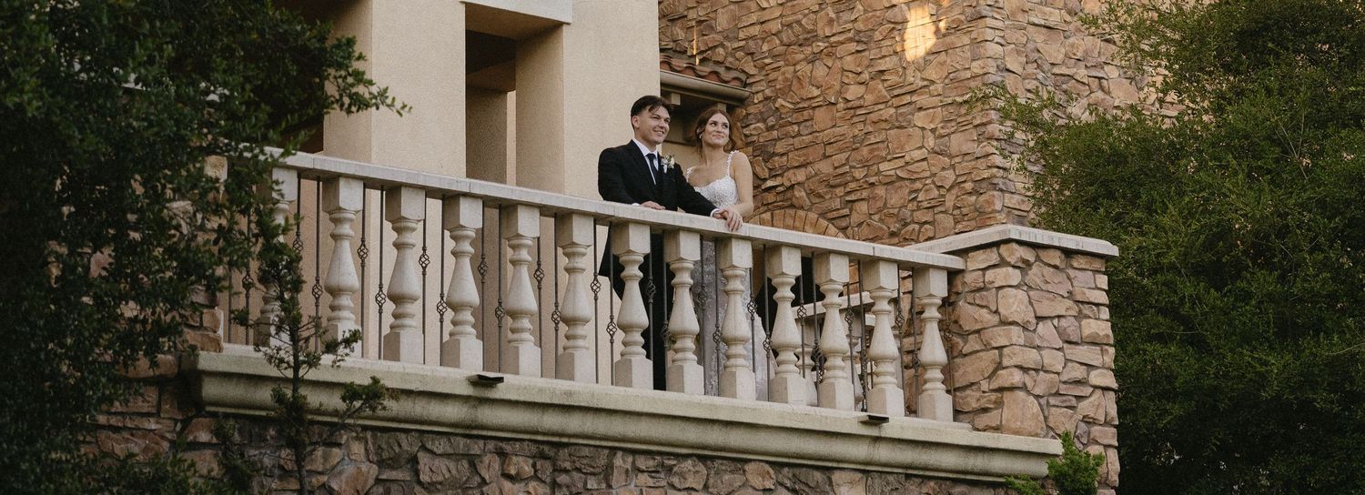 A bride and groom stand romantically on a stone balcony surrounded by lush greenery at an elegant wedding venue.