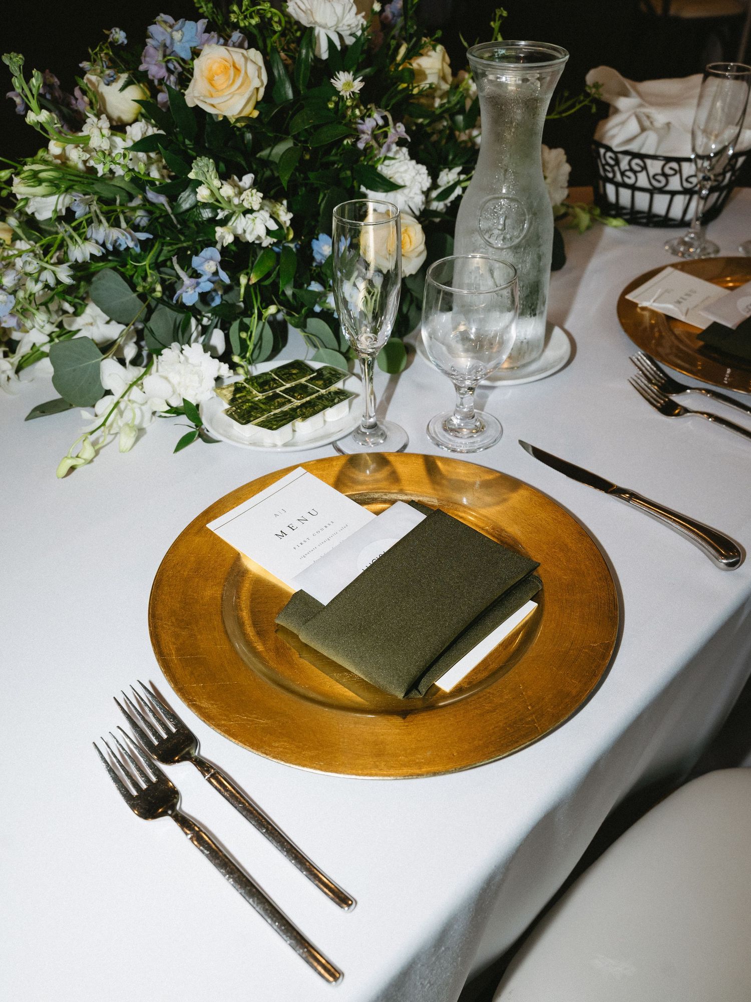 Gold charger plate with olive green napkin and menu card, flanked by gold cutlery, champagne flute, and floral centerpiece.