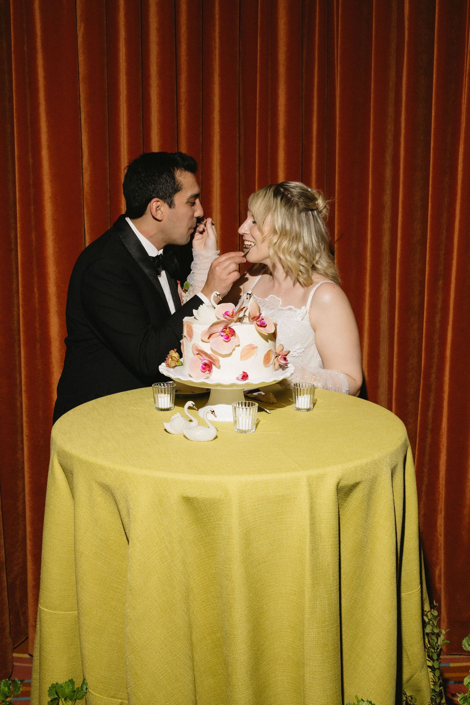 Bride and groom feeding each other wedding cake at a table with gold tablecloth against red curtain backdrop.