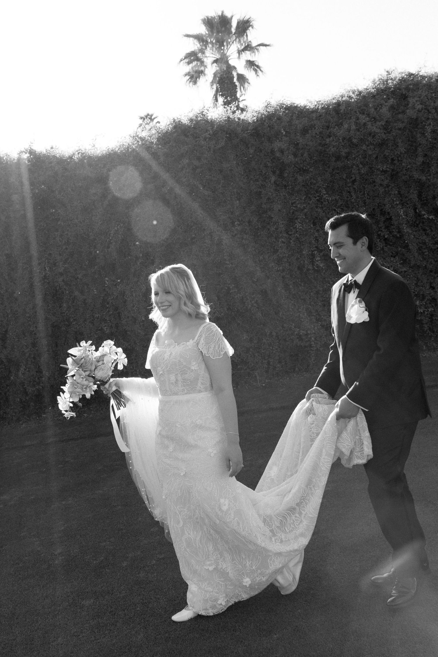 Black and white photo of bride holding bouquet and groom carrying wedding dress train outdoors.