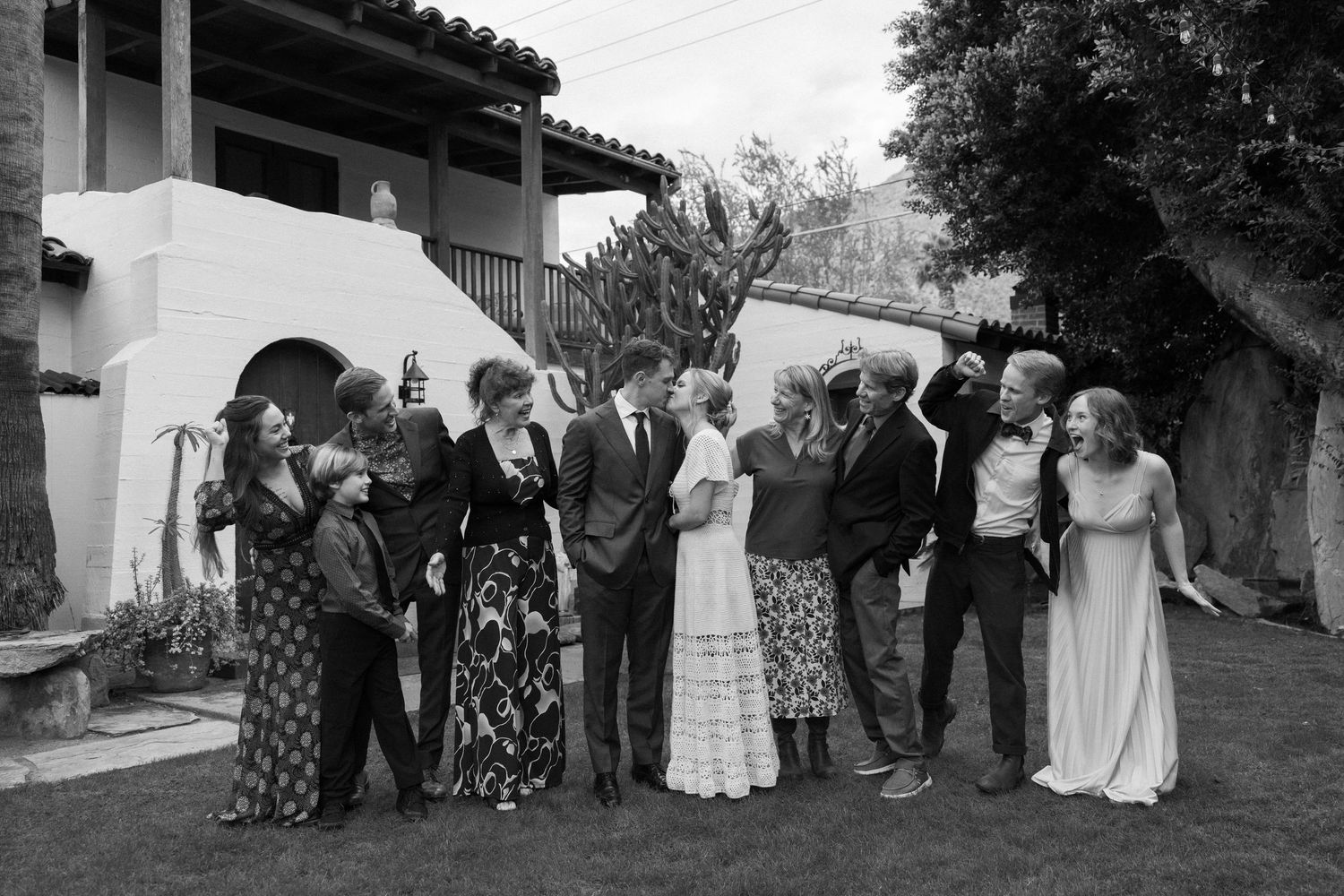 Black and white photo of a large family group laughing together outdoors at a celebration near a white building.