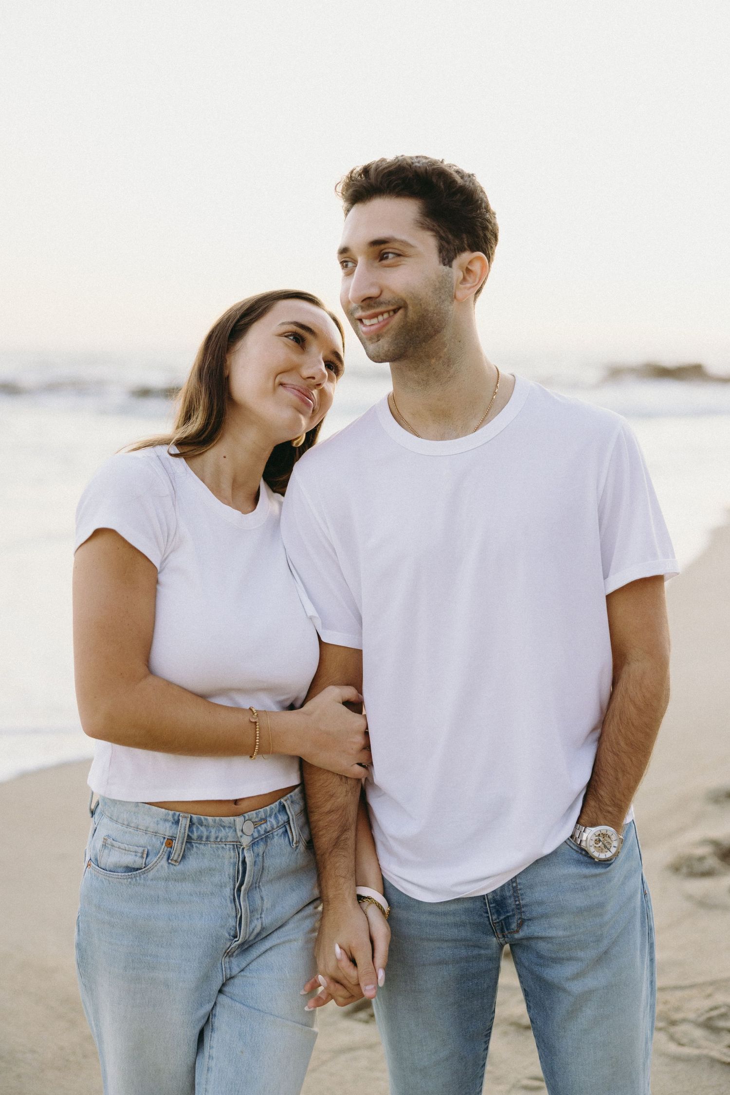 Couple in matching white t-shirts and jeans walking together on a sunny beach.