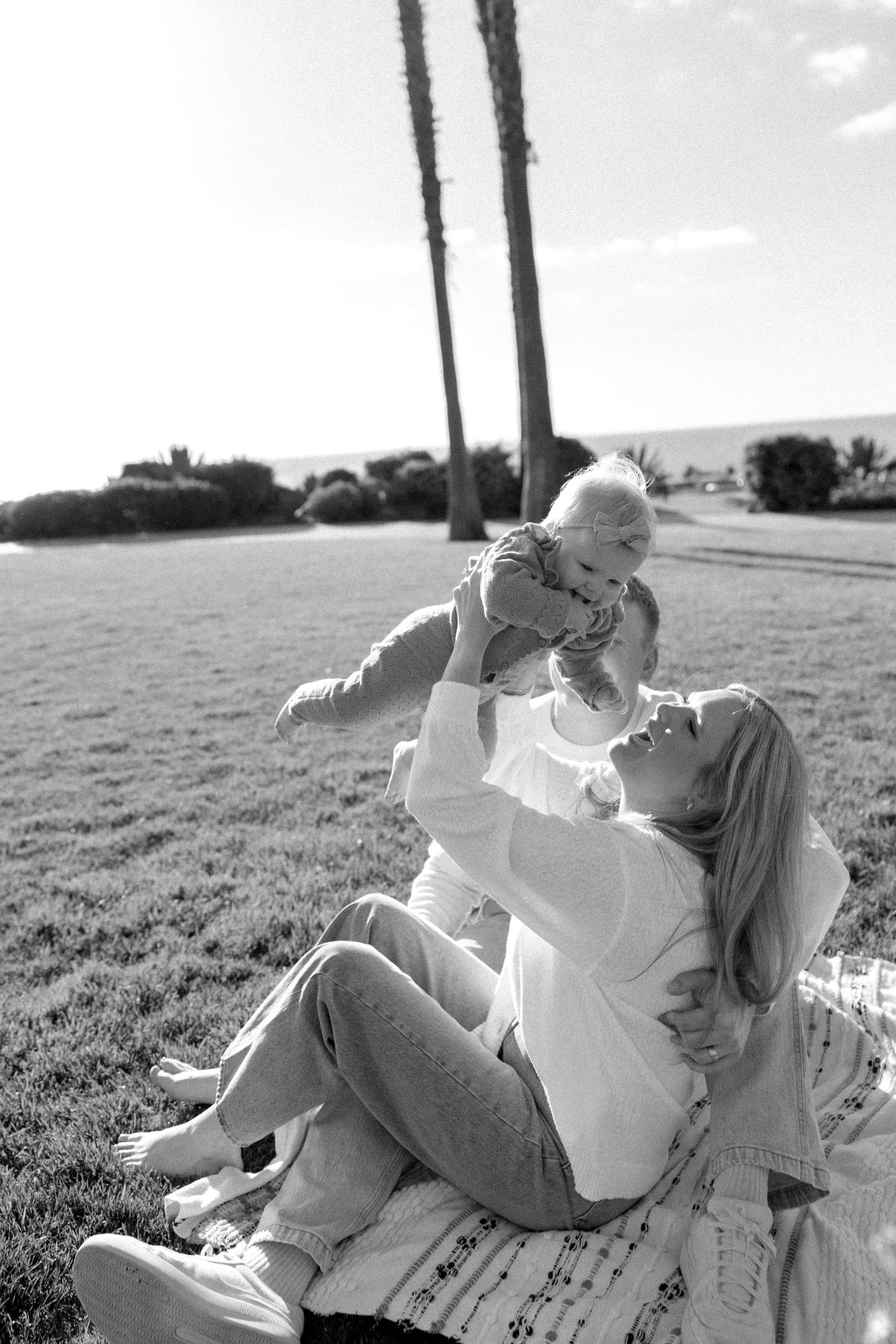 Black and white photo of couple lying in grass field with palm trees in background.