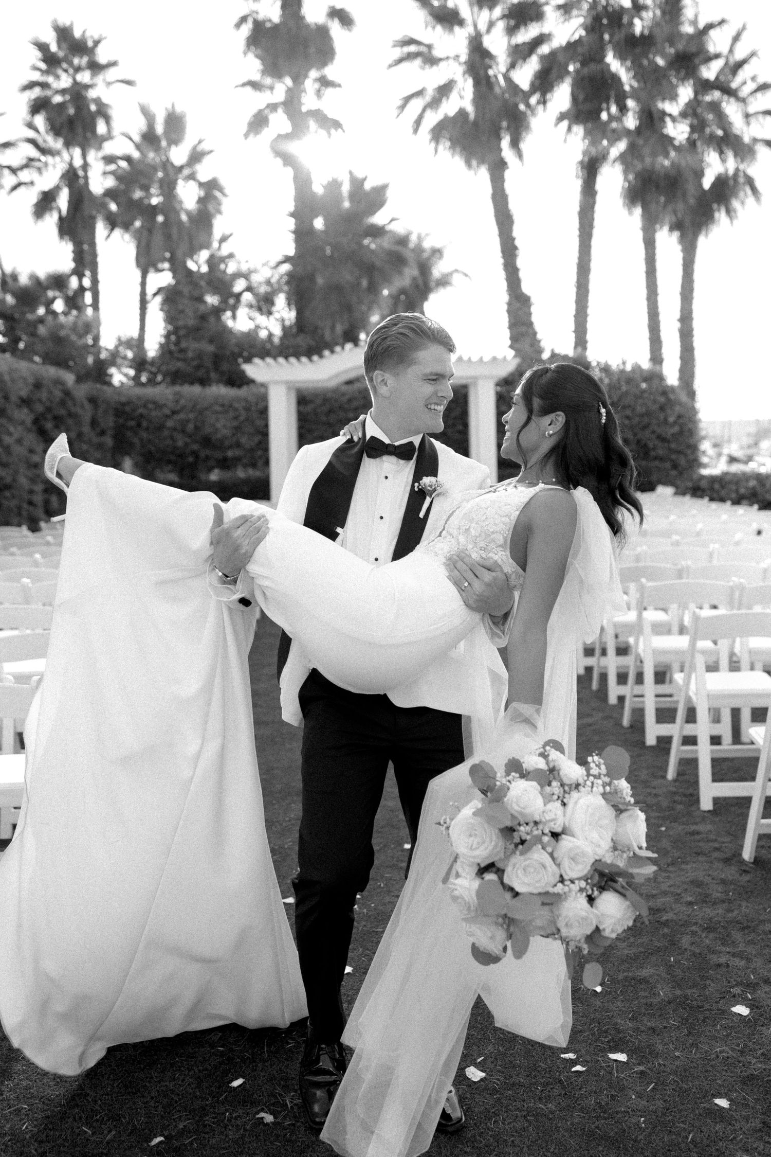 Joyful wedding couple dancing outdoors with palm trees and white chairs in background.