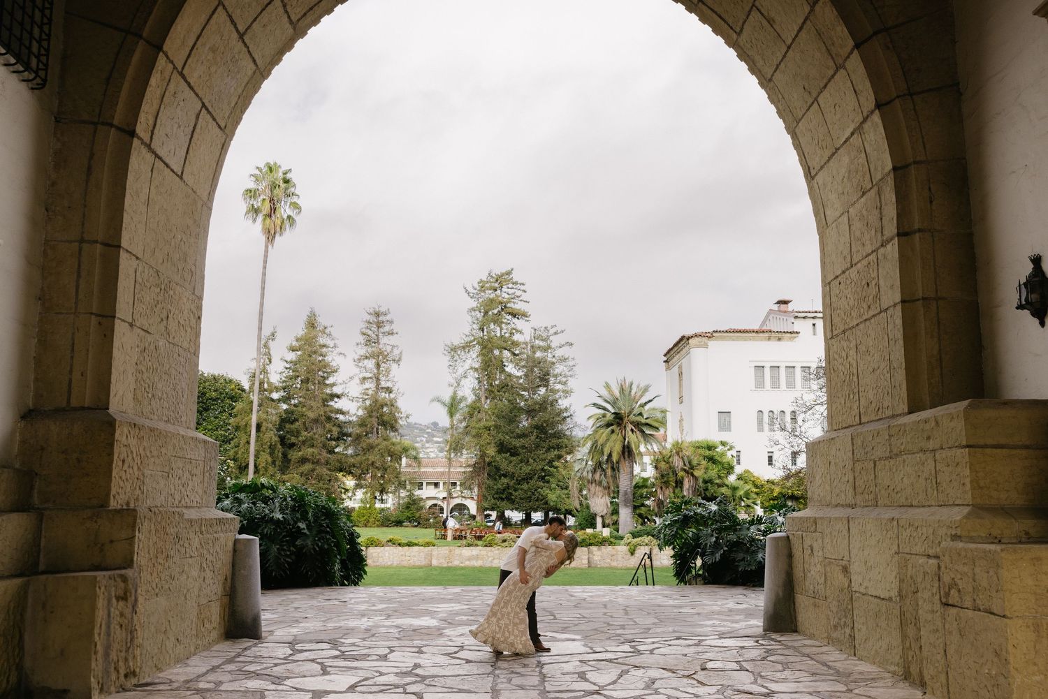 Stone archways frame a view of a white Spanish colonial building and palm trees in the distance.