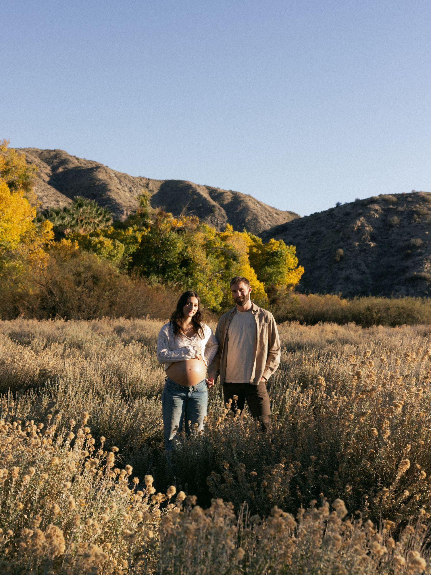 A couple stands in a field of sagebrush with mountains and fall colors behind them.