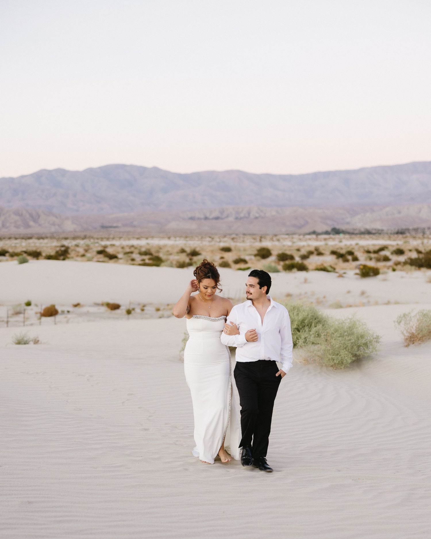 Wedding couple walks together across desert sand dunes during sunset photoshoot.