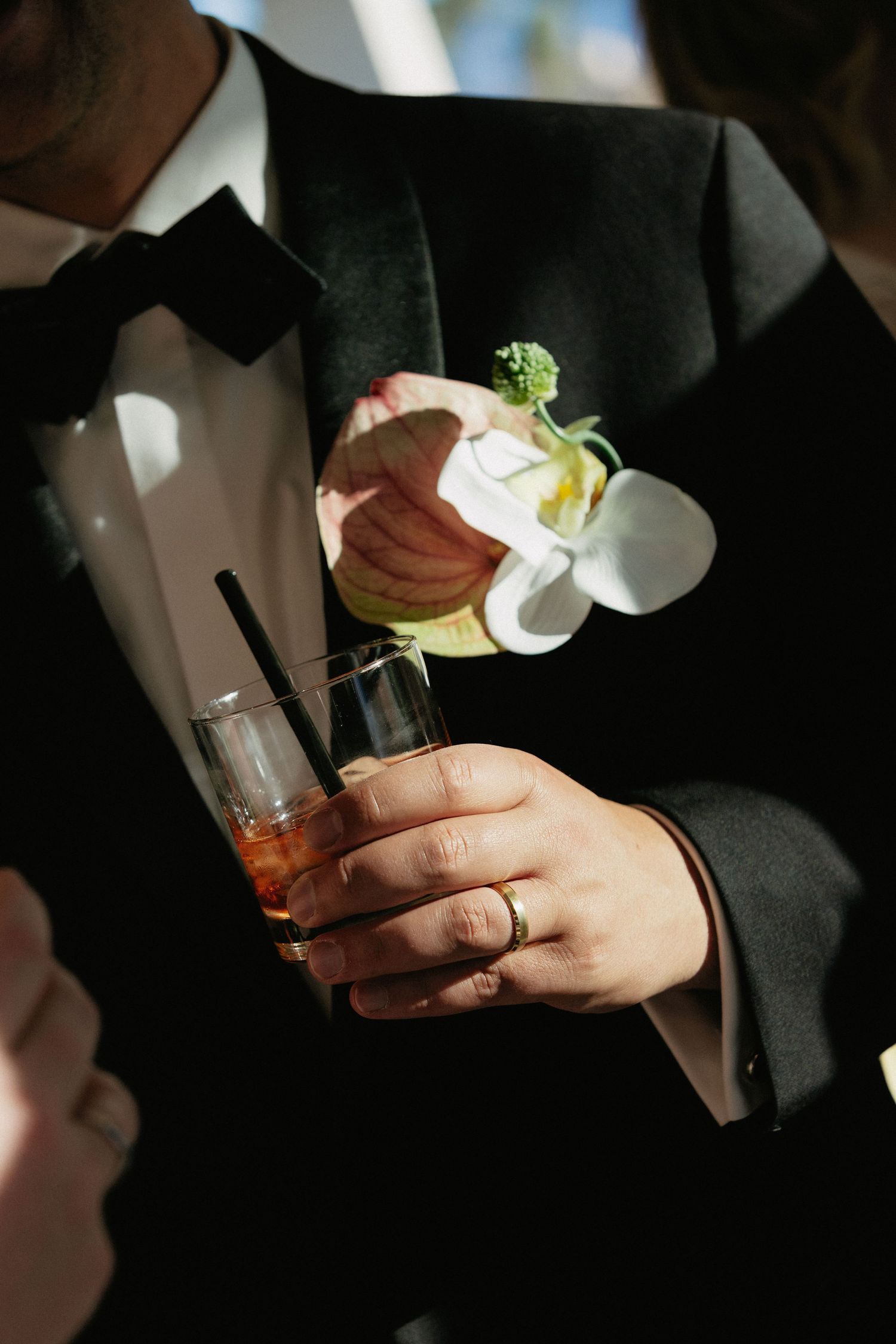 Groom in black tuxedo holding whiskey cocktail with floral boutonniere featuring white orchid and green accents.