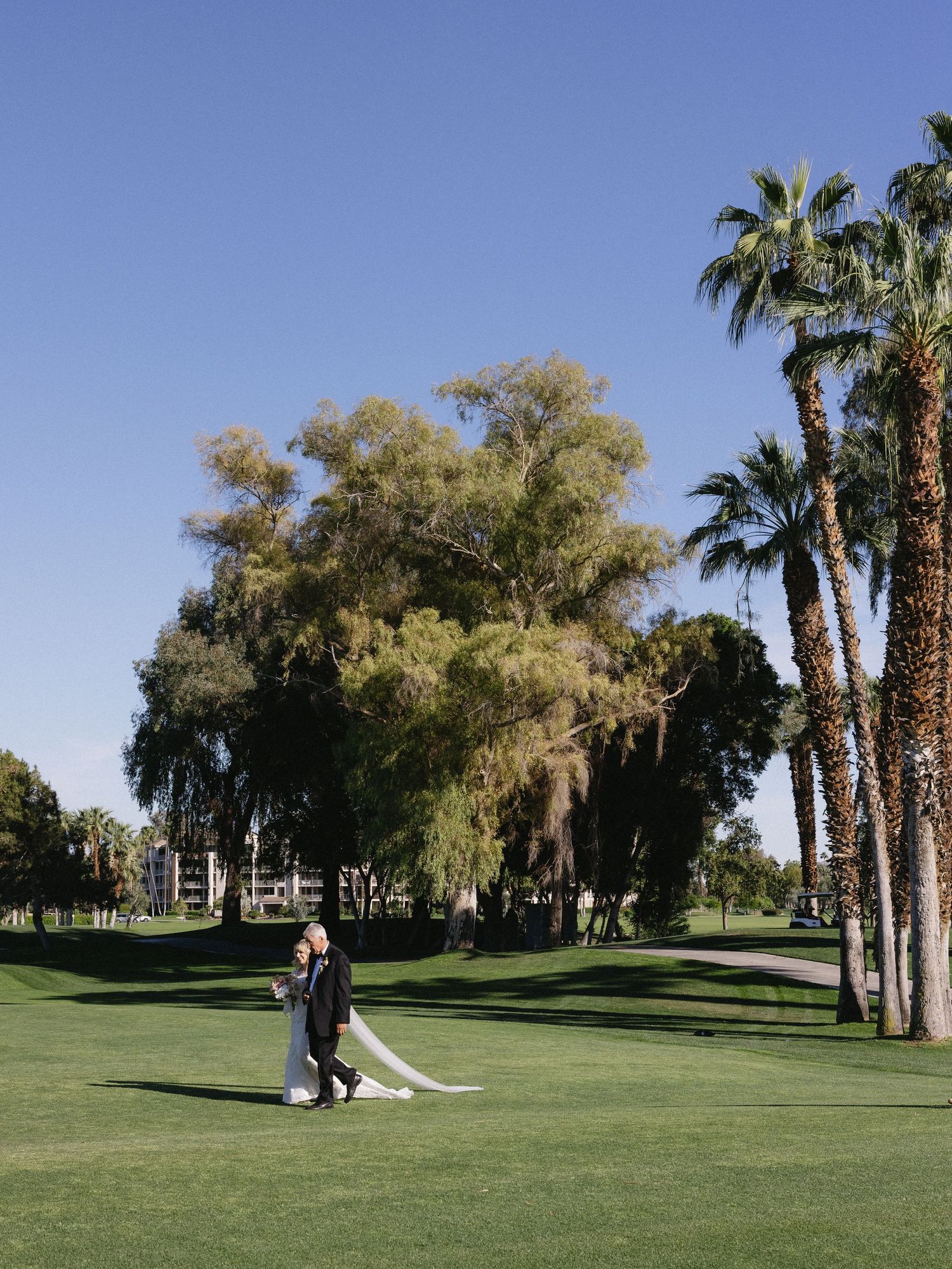 Bride and groom embrace on lush green golf course surrounded by palm trees under a clear blue sky.