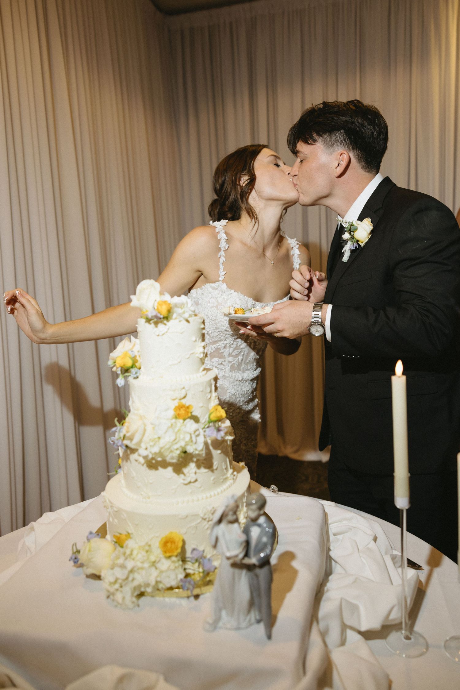 Bride and groom kissing while cutting their white wedding cake decorated with yellow roses and figurines.