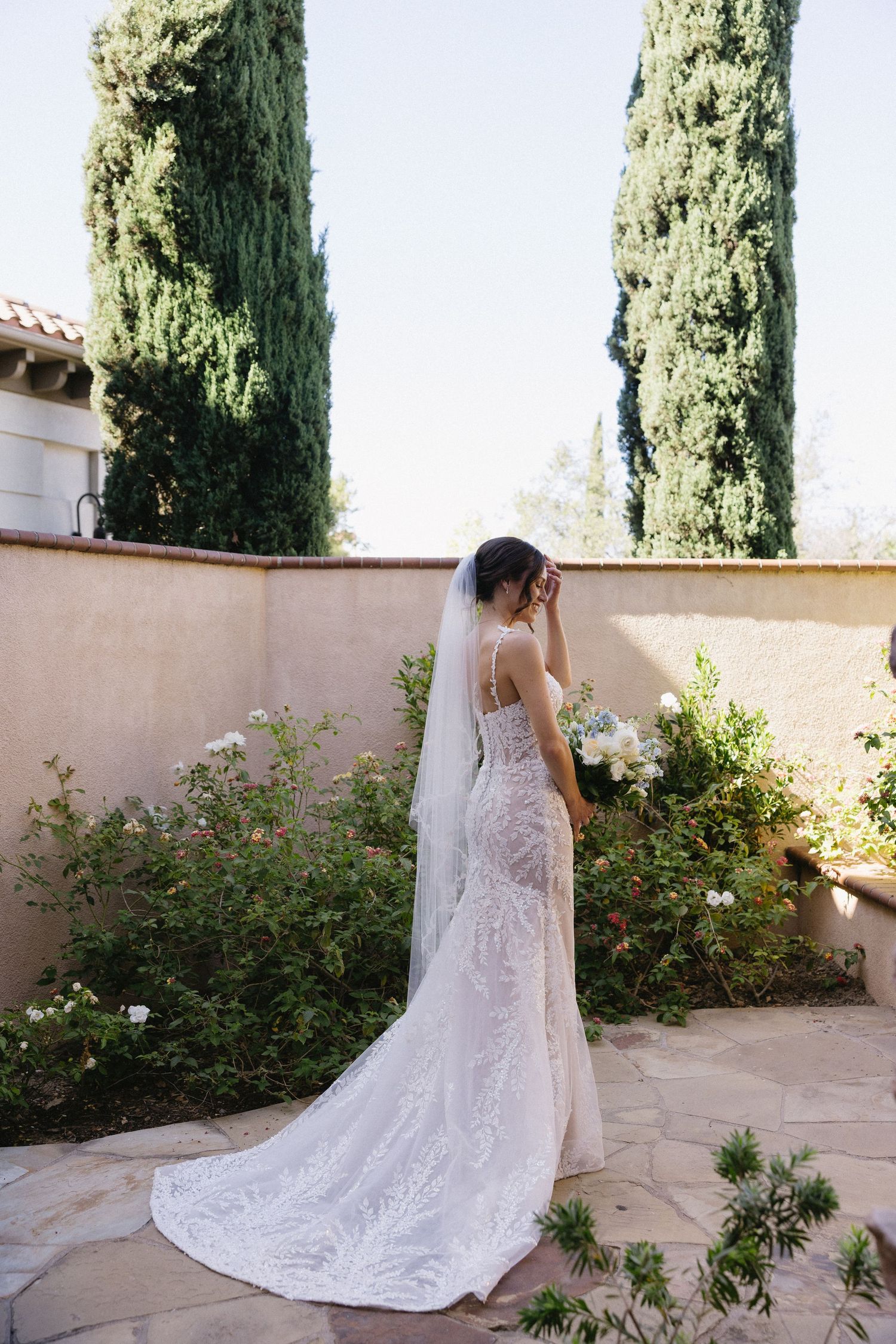 Bride in lace wedding gown with veil holding bouquet in garden courtyard with cypress trees.