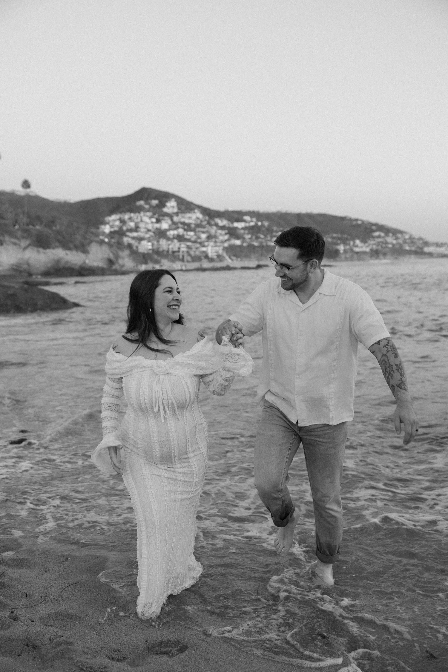 A couple plays in the shallow ocean water with coastal mountains in the background.