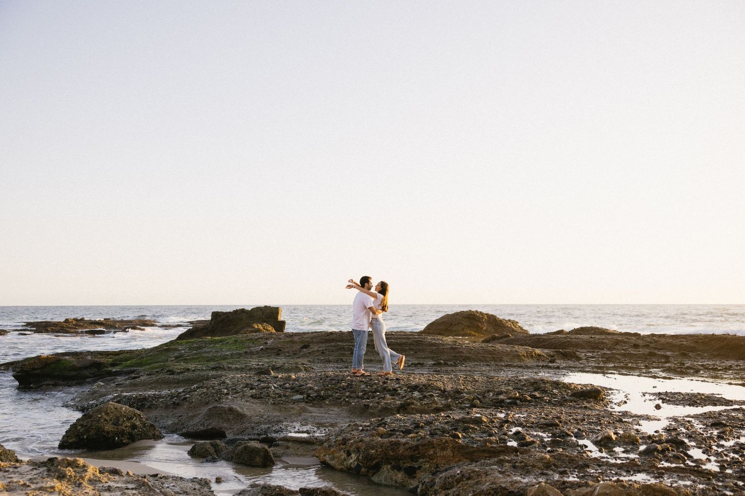 Couple walking along a rocky beach coastline at sunset with waves crashing against the shore.