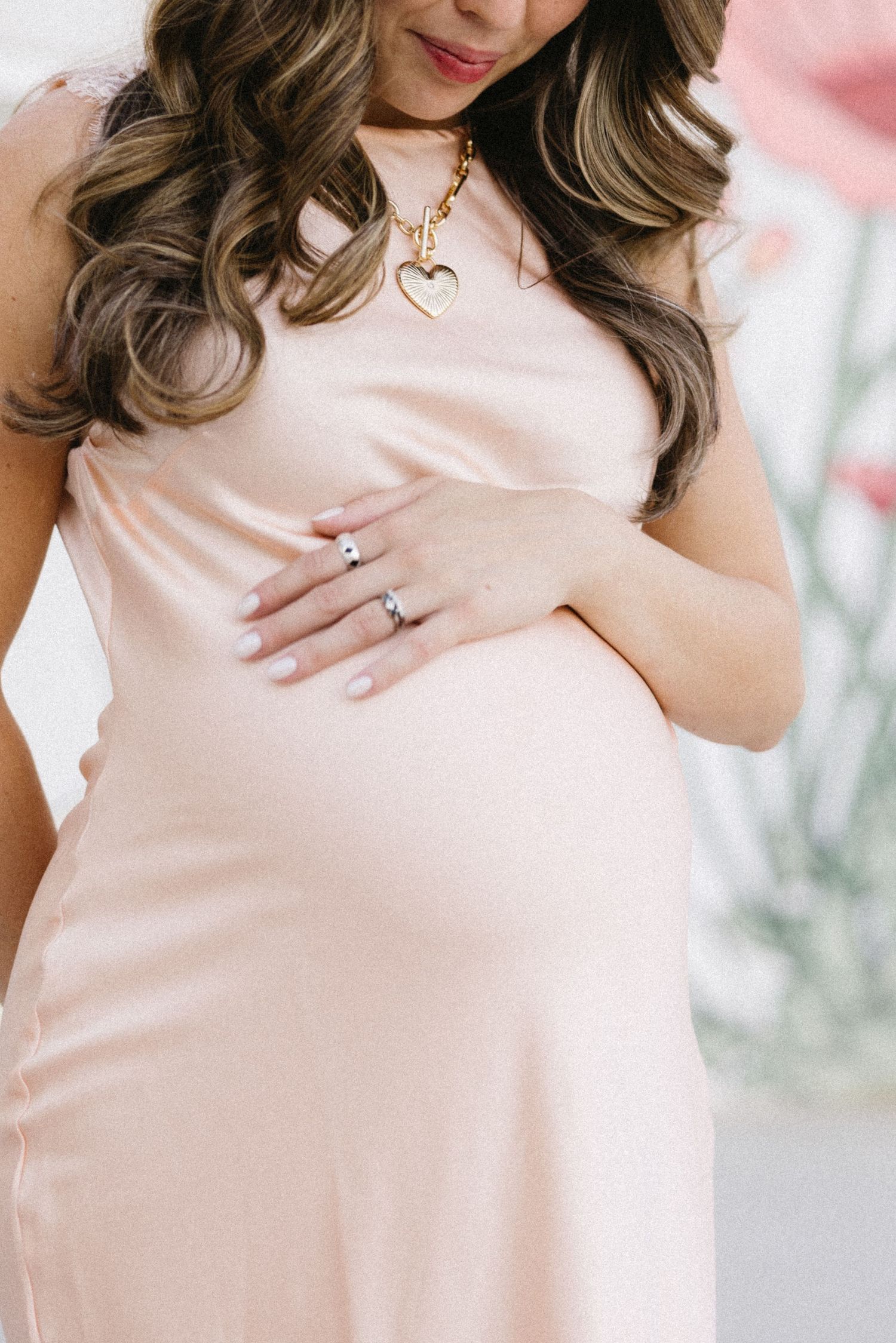 Close-up maternity details with heart necklace and pink dress against floral backdrop.