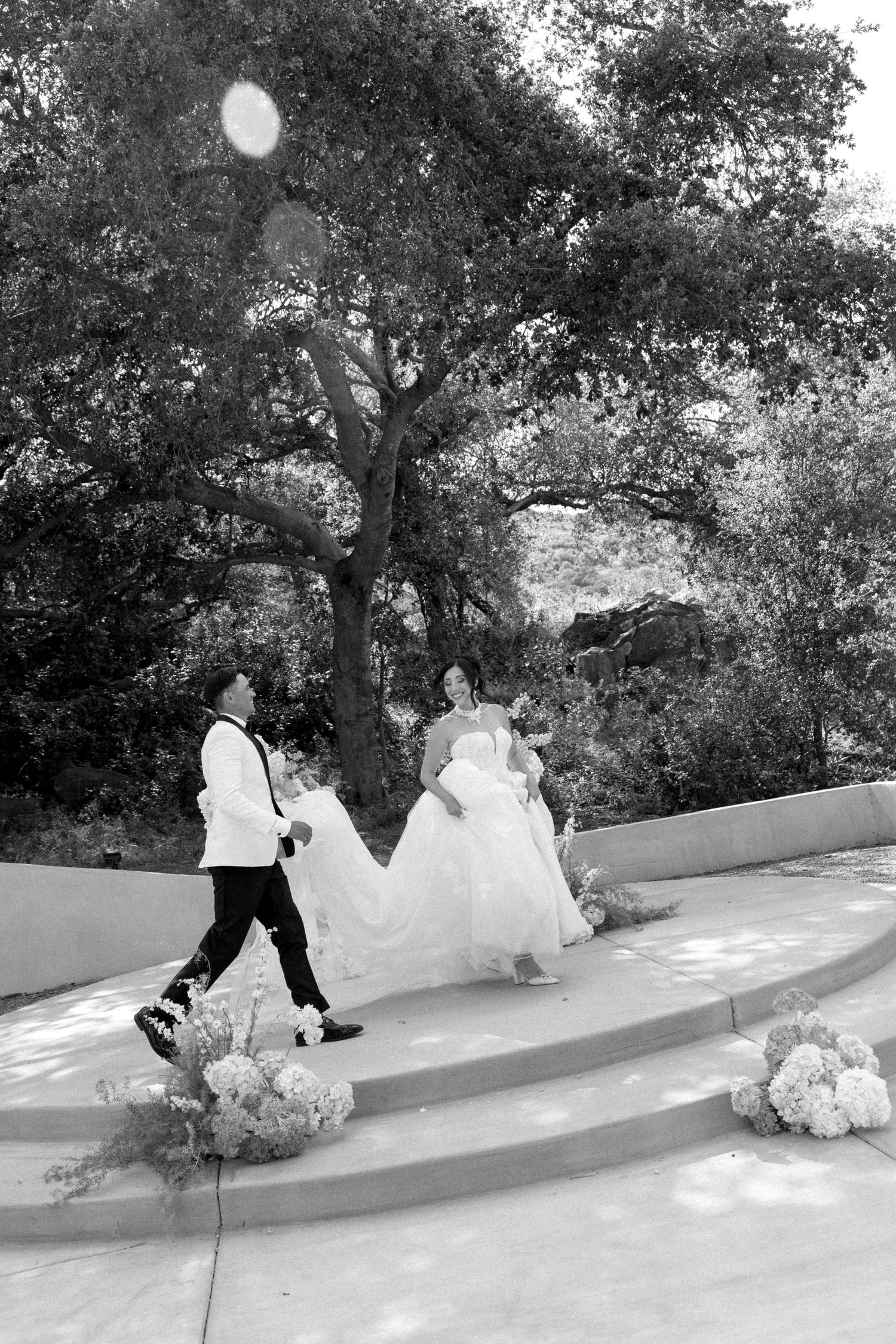 Black and white photo of a bride and groom walking down outdoor steps surrounded by floral arrangements and trees.