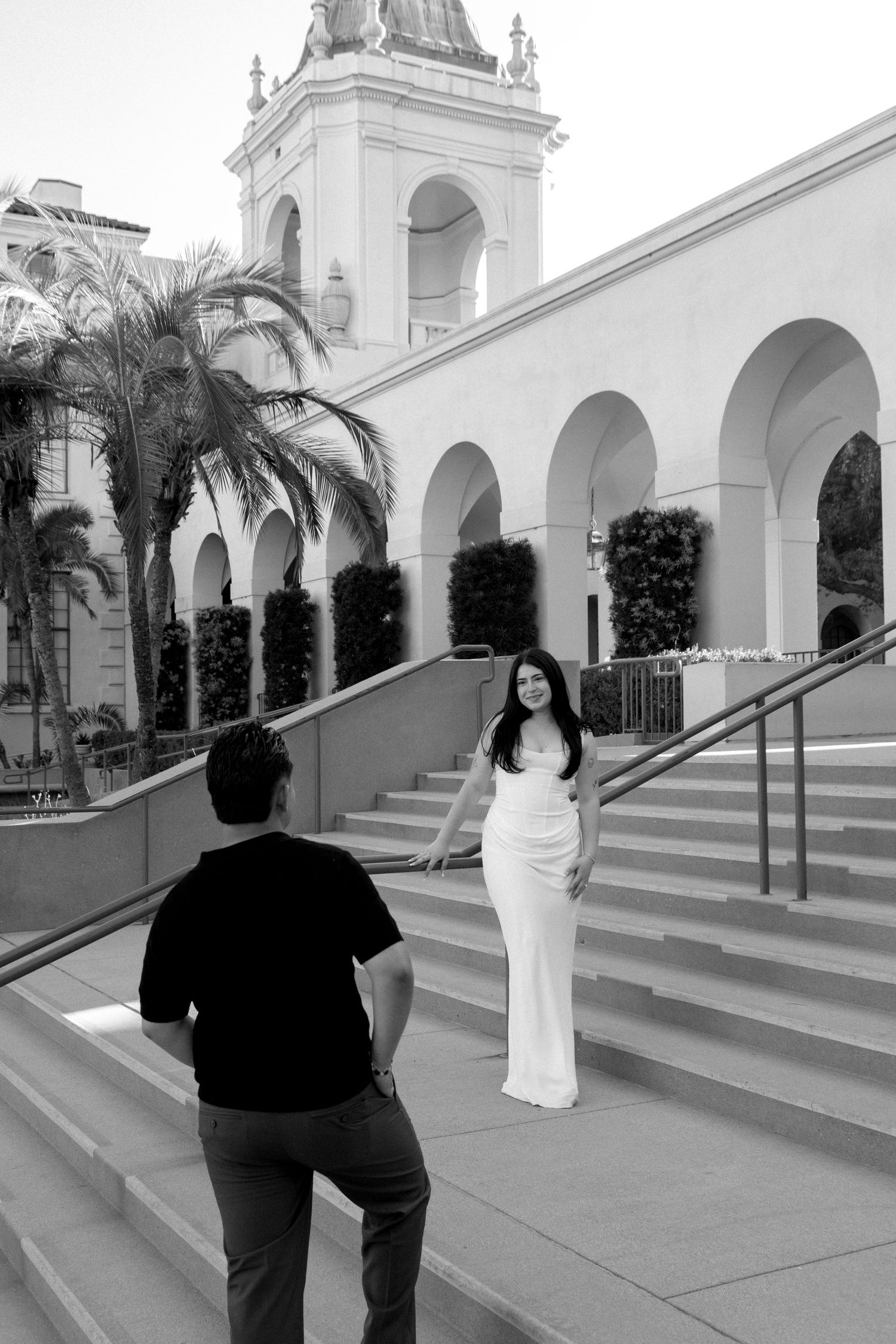 Black and white photo of a woman in a white gown posing on steps outside an elegant building with arches and a tower.