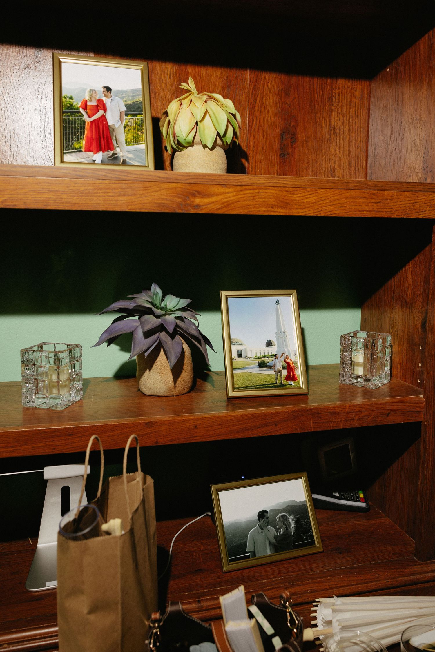 Wooden bookshelf decorated with framed couple photos, succulent plants, and glass candle holders against a green wall.