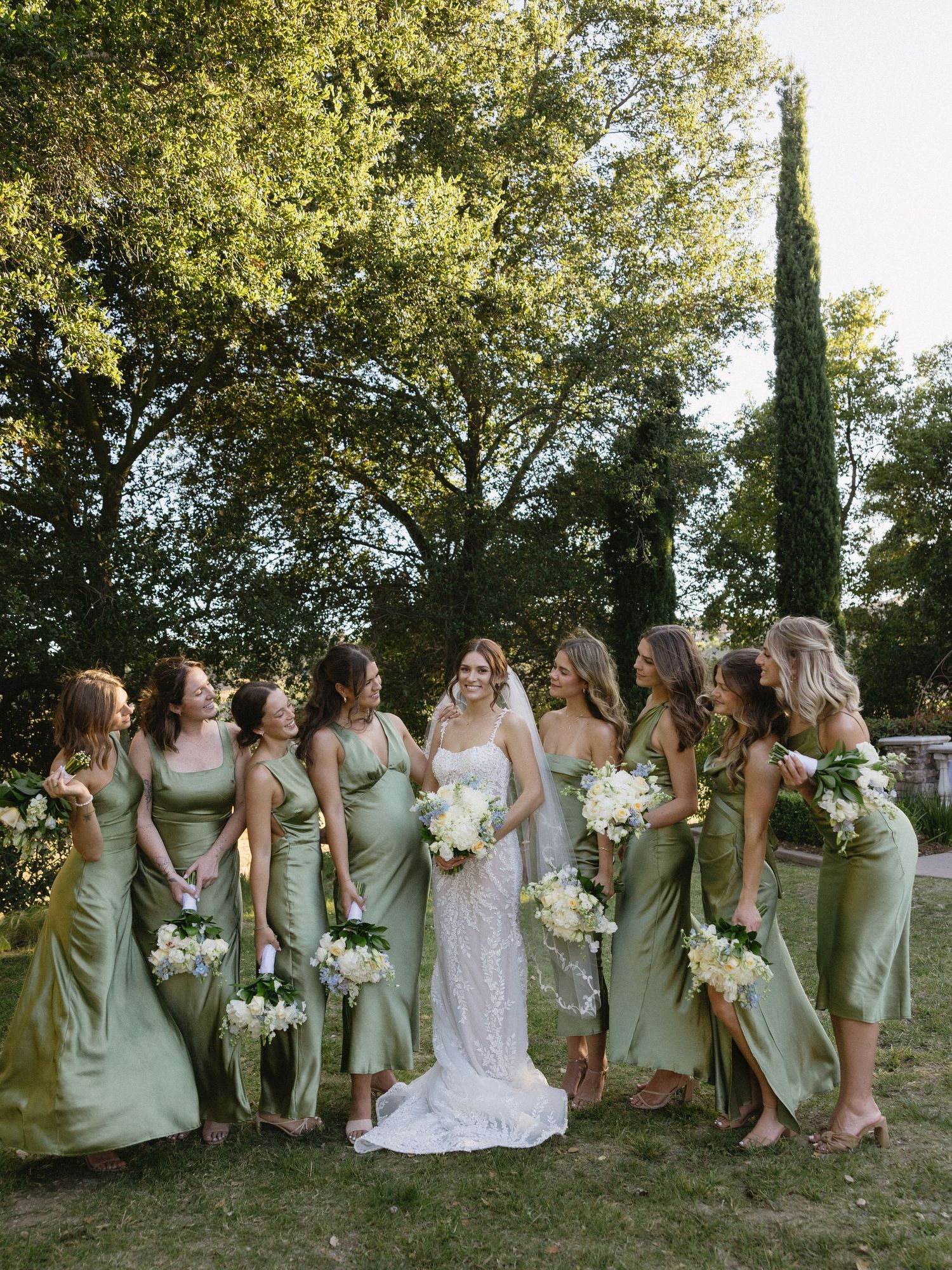 Bride in white lace gown stands with bridesmaids in sage green satin dresses holding white bouquets outdoors.