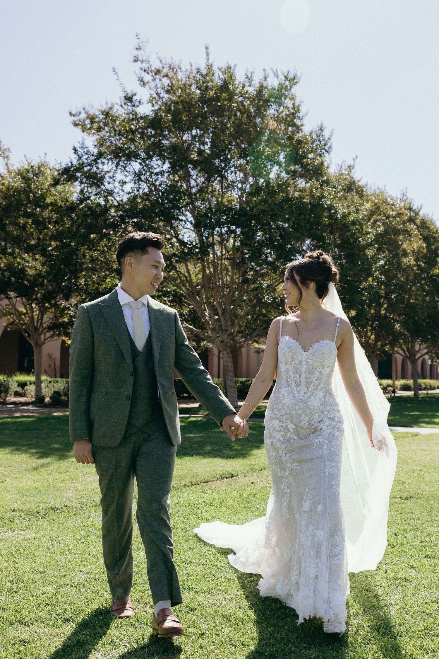 A bride and groom hold hands walking on green grass outdoors on their wedding day in elegant attire.