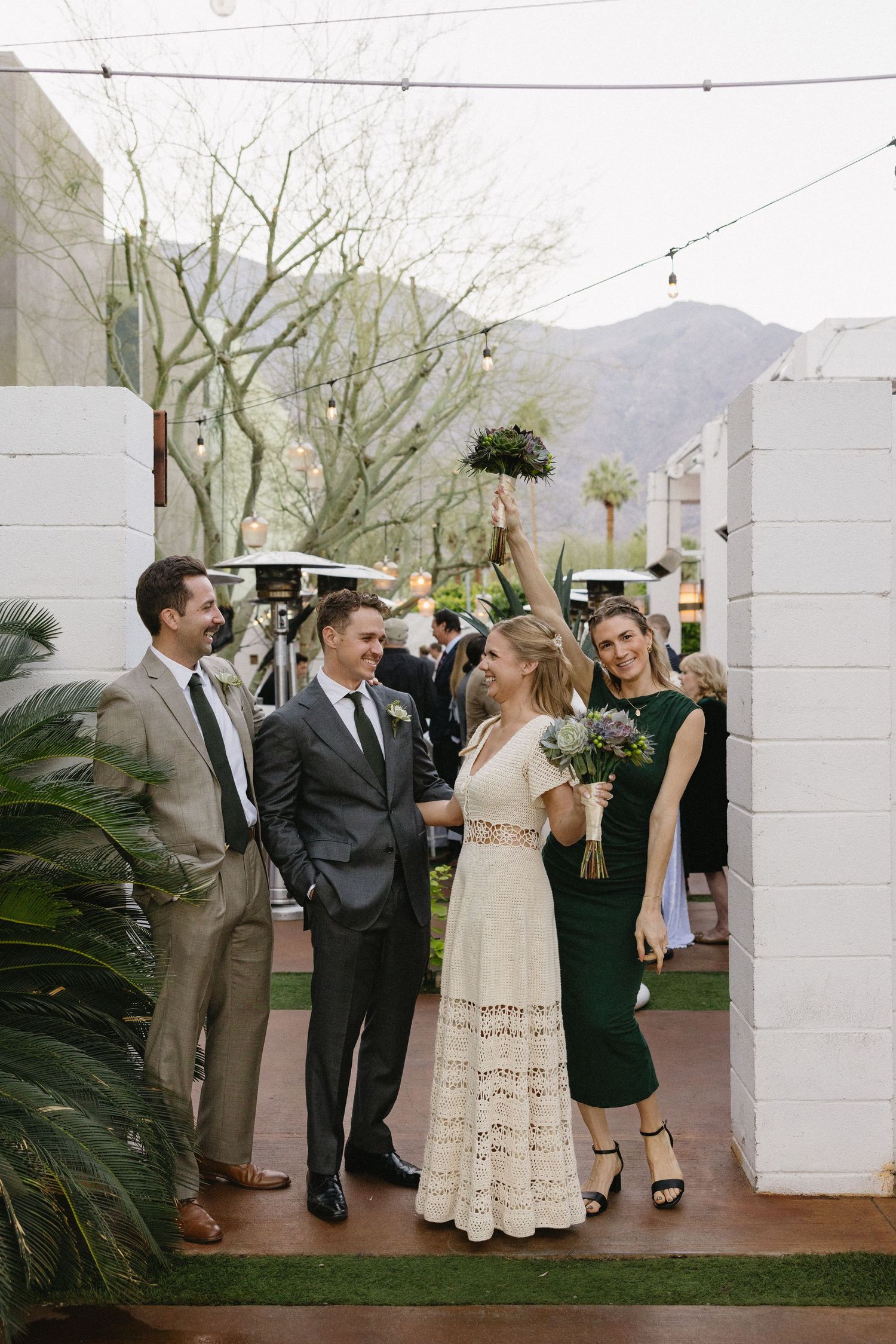 Wedding party celebrating outdoors in Palm Springs, bride in lace dress holding bouquet aloft with guests in formal attire.