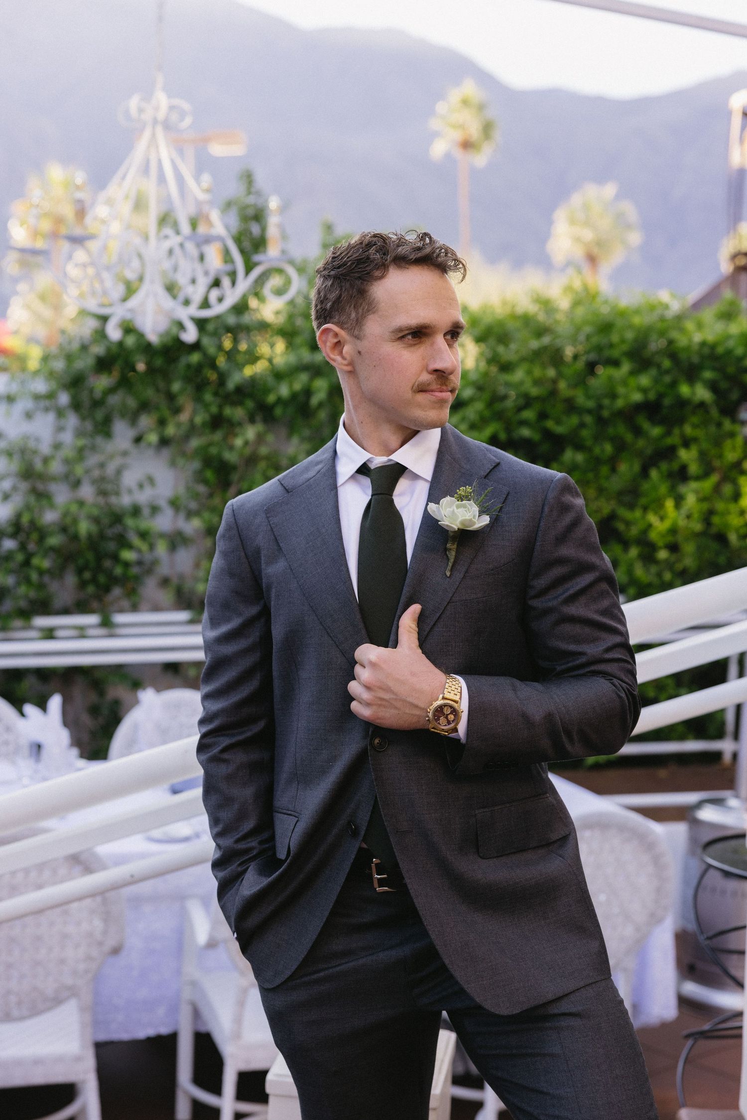 Groom in dark suit with boutonniere and gold watch poses outdoors at elegant wedding venue with palm trees.