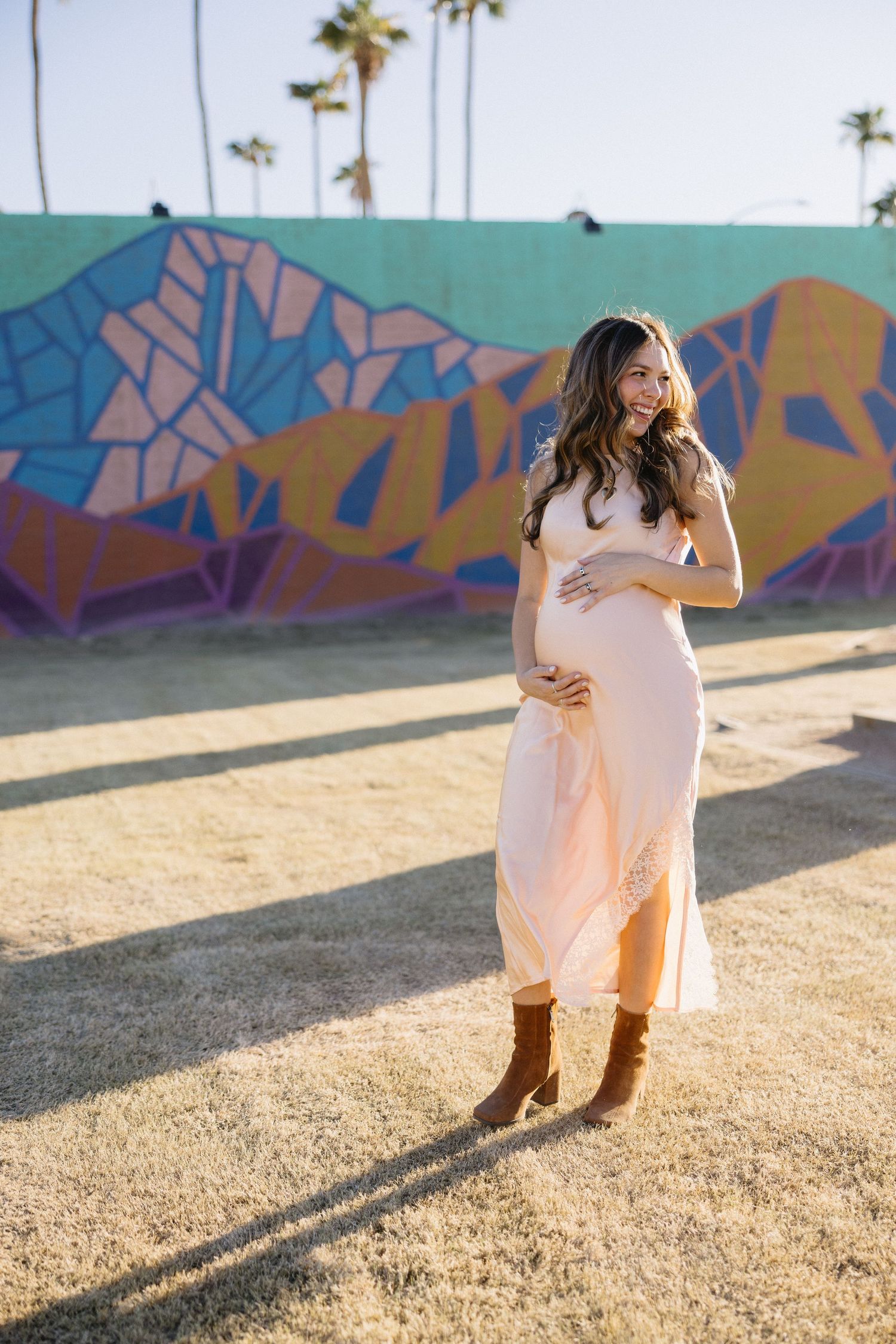 Expectant mother in pink dress poses near colorful geometric street art during sunset.