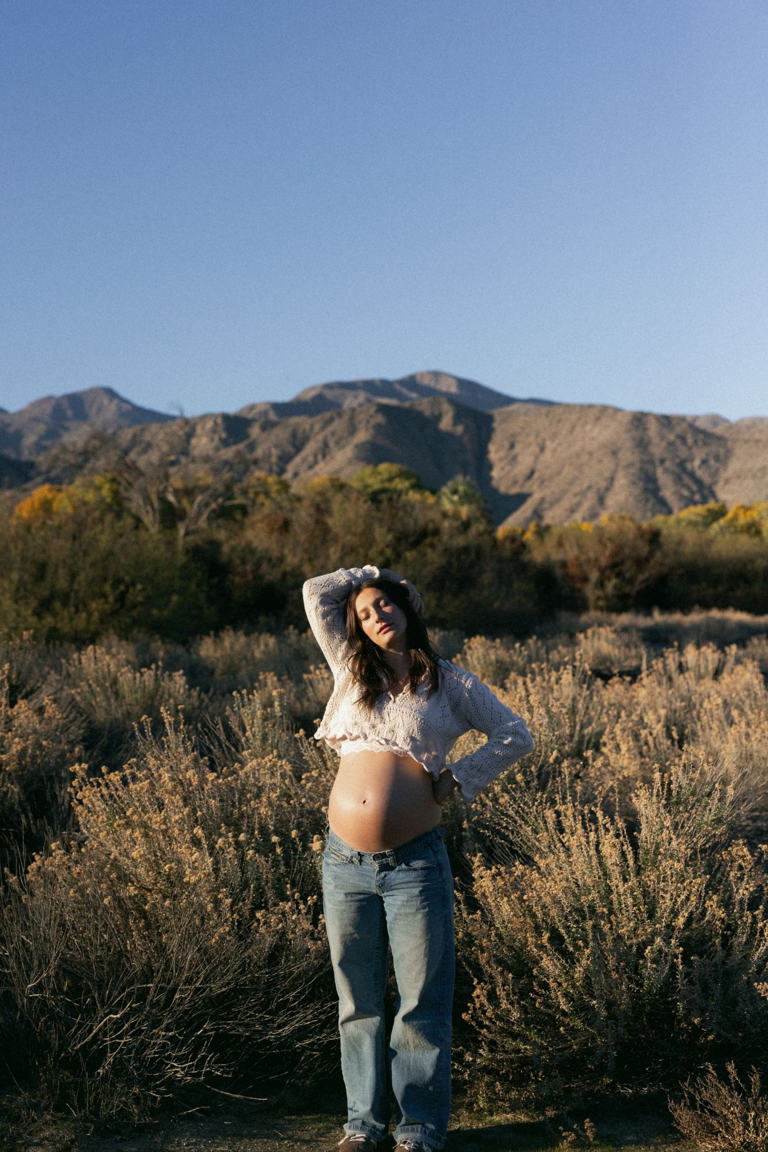A maternity photo in a desert landscape with mountains and autumn foliage in the background.