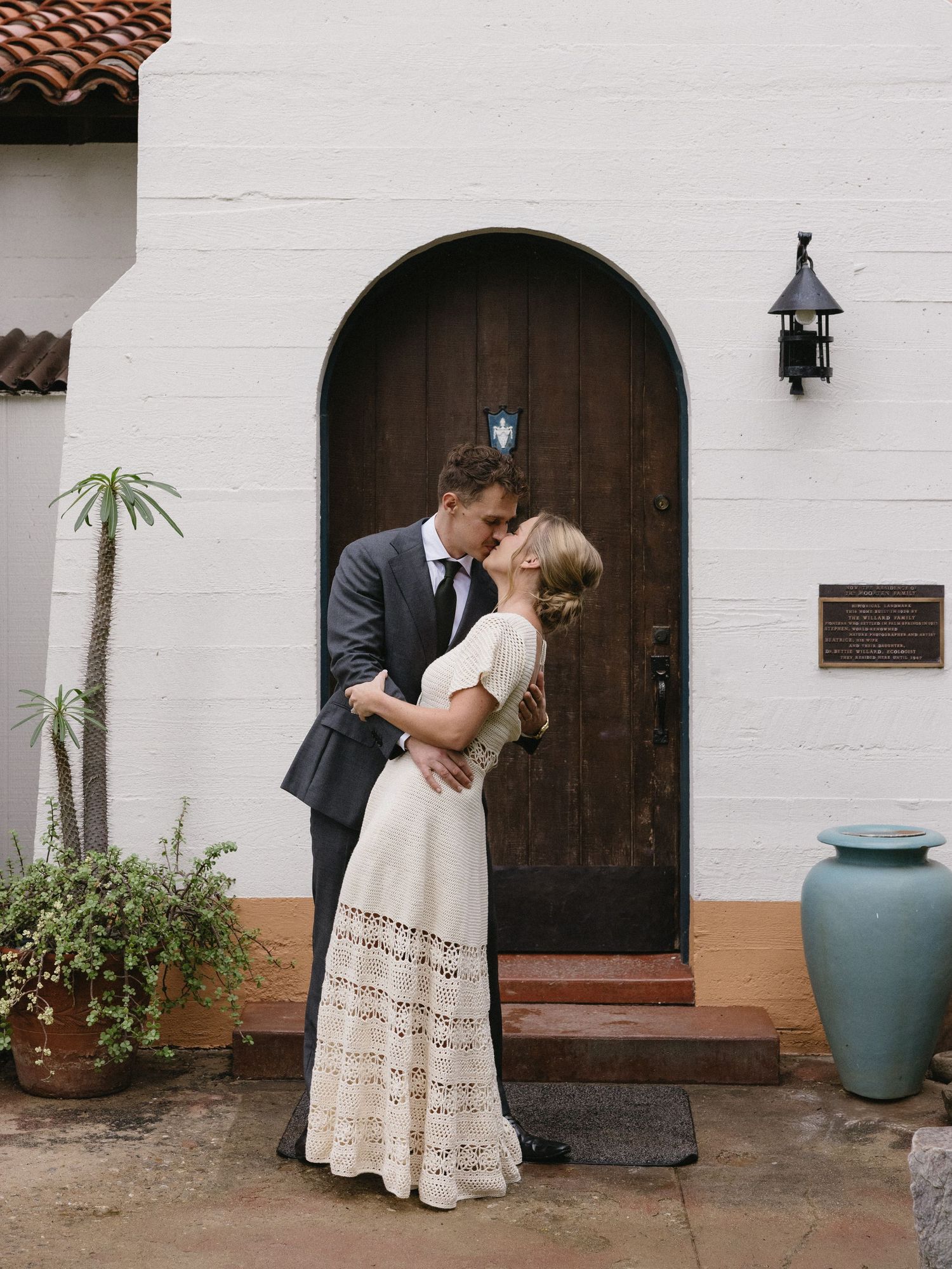 Couple sharing a romantic kiss in wedding attire outside an arched wooden door of a white stucco building.
