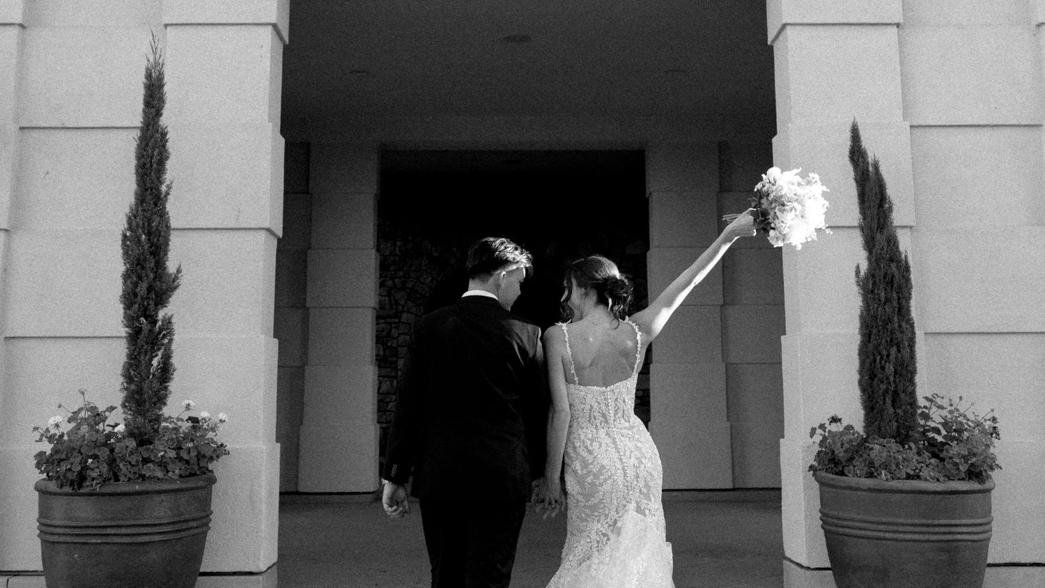 Black and white photo of bride and groom entering wedding venue, bride holding bouquet aloft.