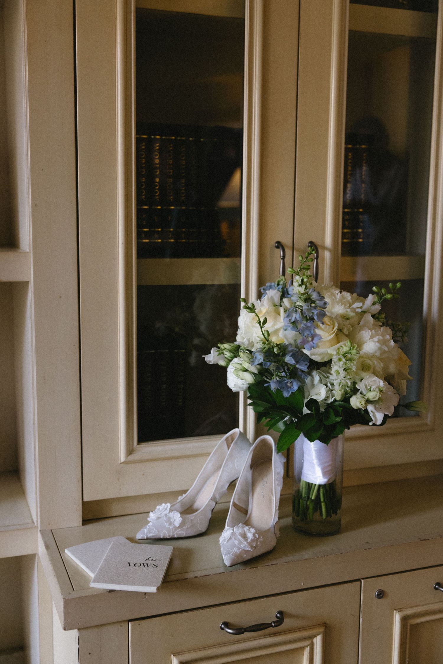 White bridal heels and bouquet of white and blue flowers on a cream cabinet with glass doors and books.