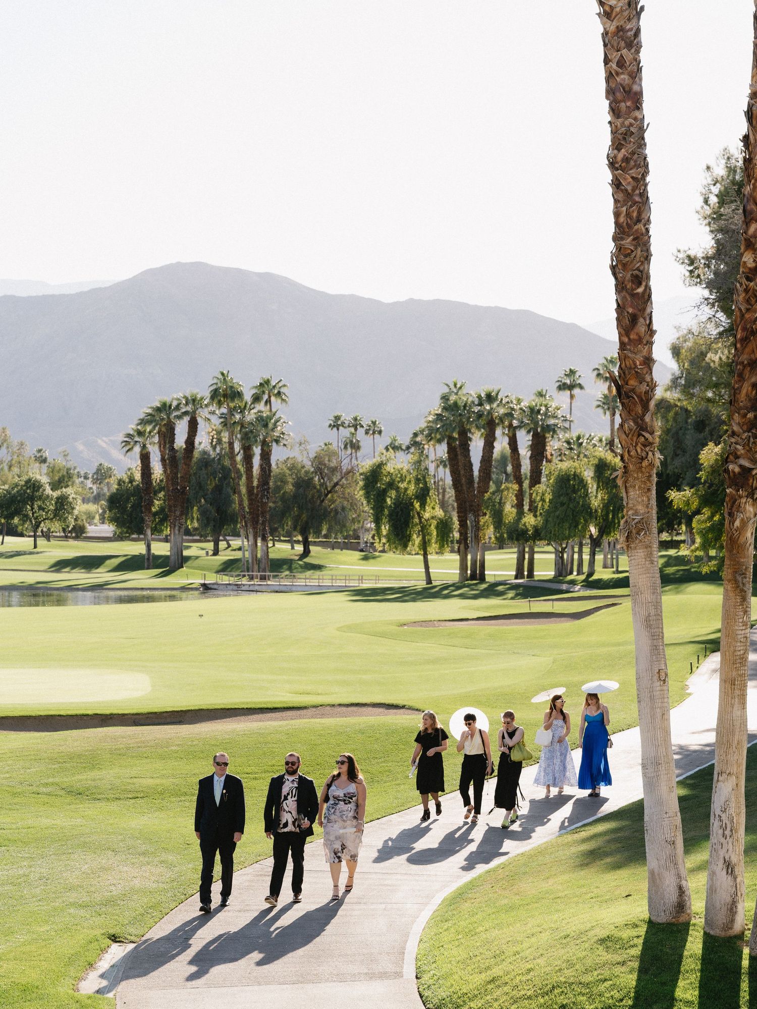 Guests walking along a path at a lush golf course with palm trees and mountain views on a sunny day.
