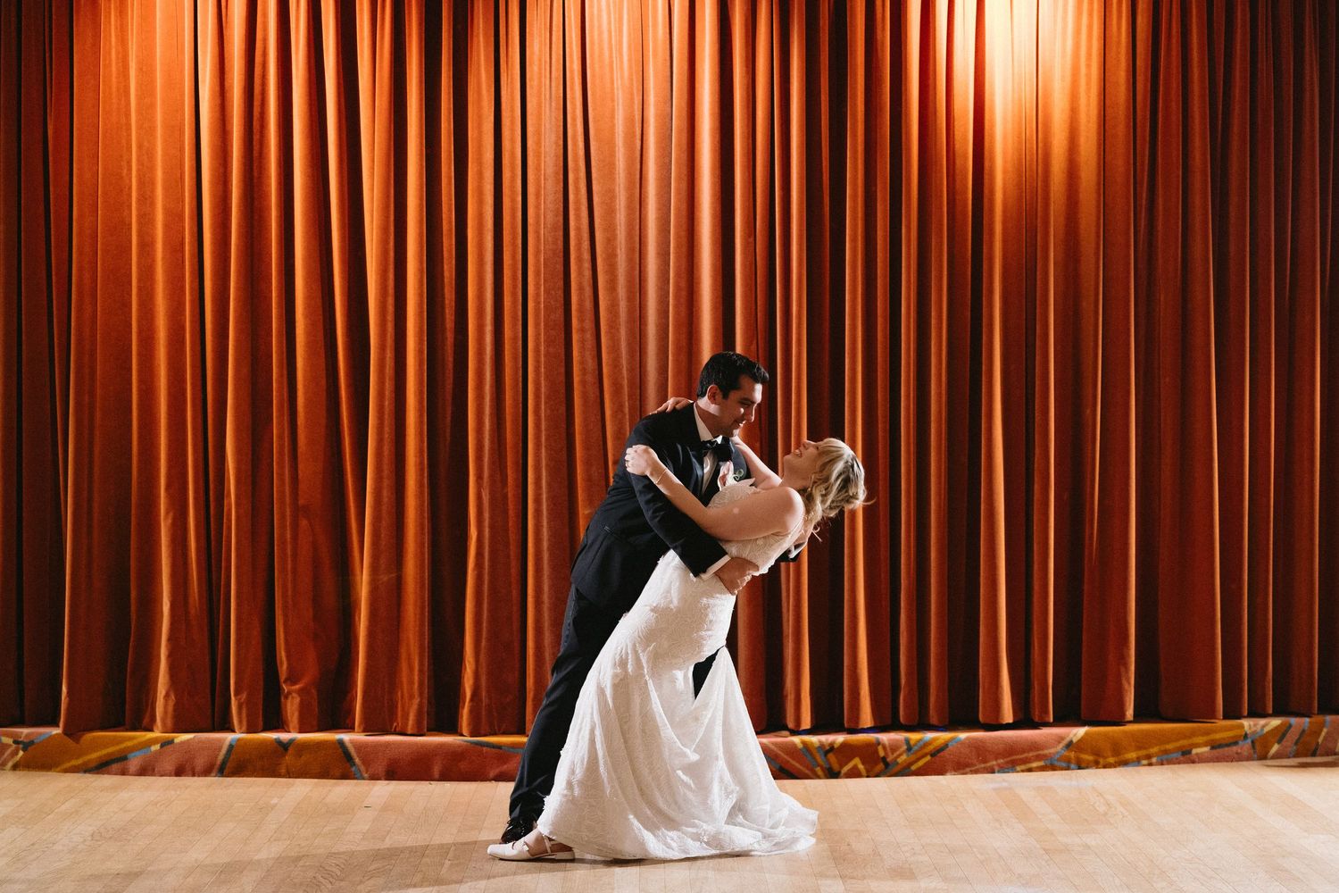 Bride and groom share romantic first dance dip in front of elegant red stage curtains at wedding reception.