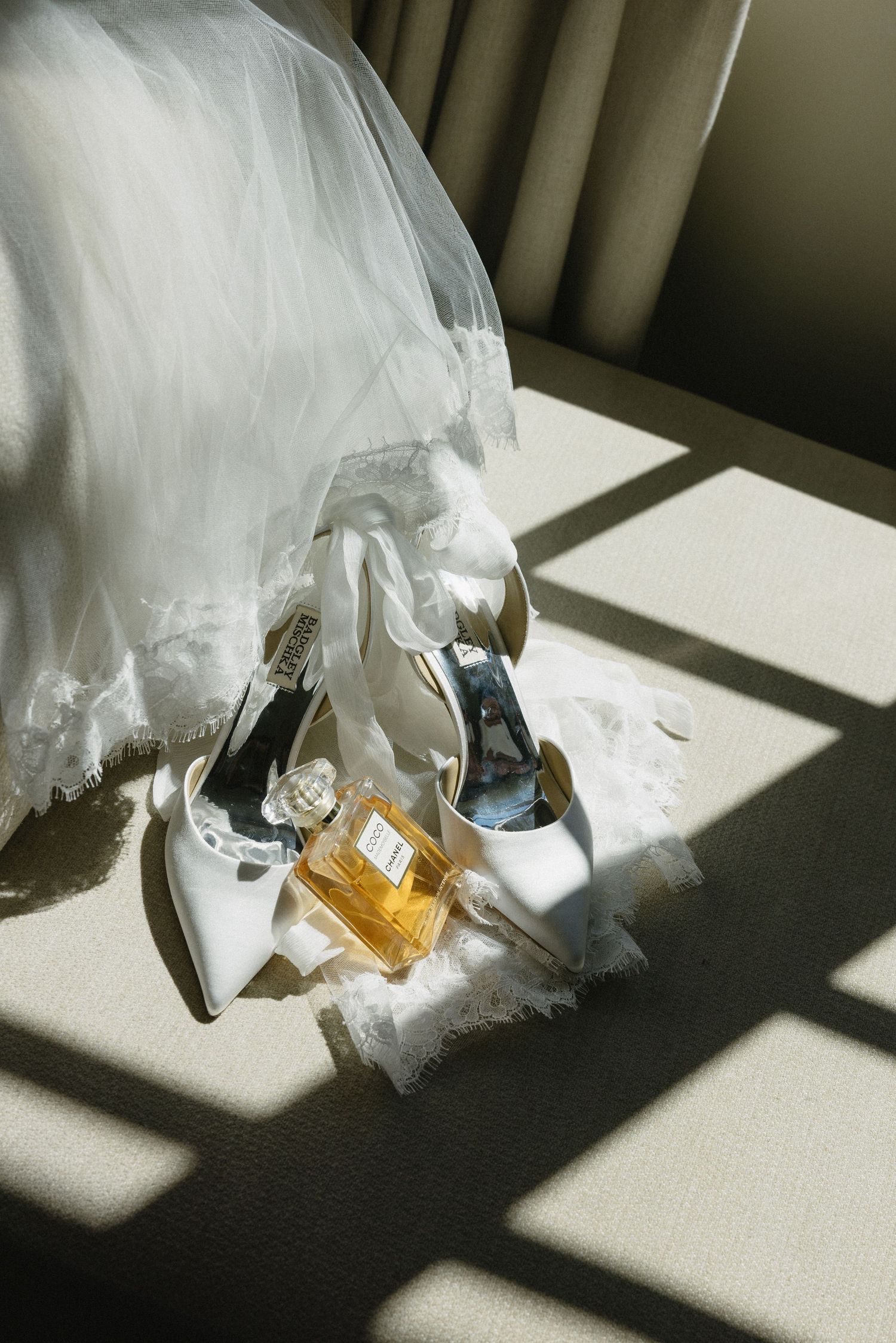 White bridal heels and Chanel Coco perfume bottle arranged with wedding veil in warm sunlight on neutral surface.