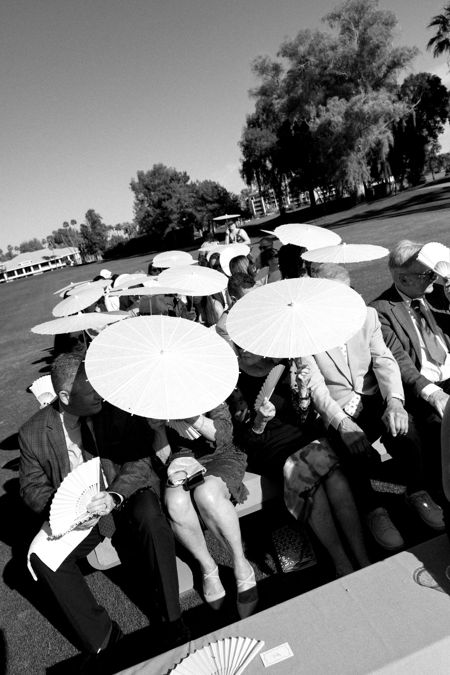 Black and white photo of wedding guests shielding from sun with white parasols during an outdoor ceremony.