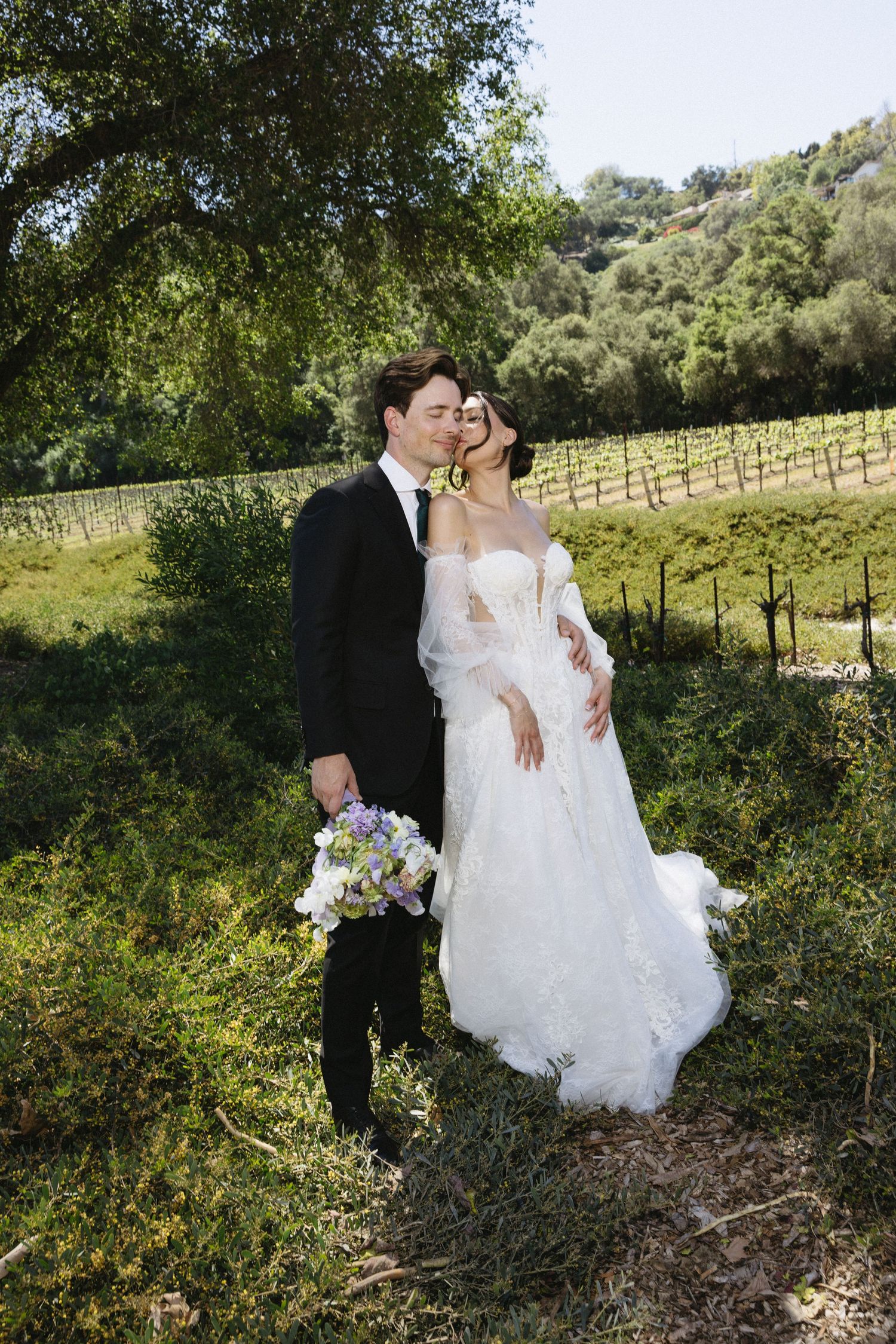 Bride and groom embrace in a vineyard setting, she in lace gown, he in black tux holding a purple bouquet.