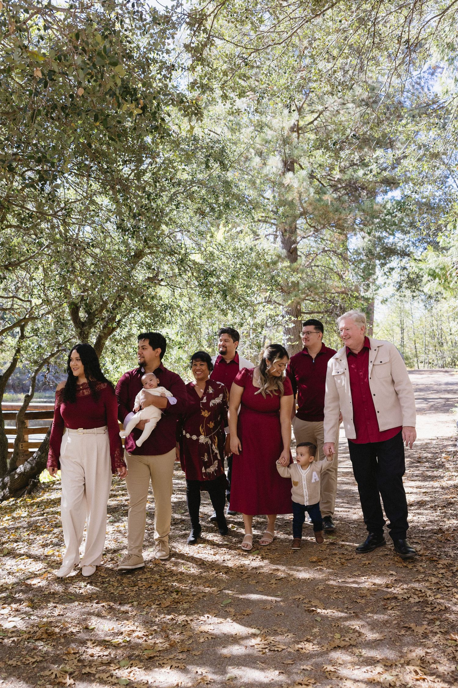 Large family group wearing burgundy and white outfits posing under shady trees.
