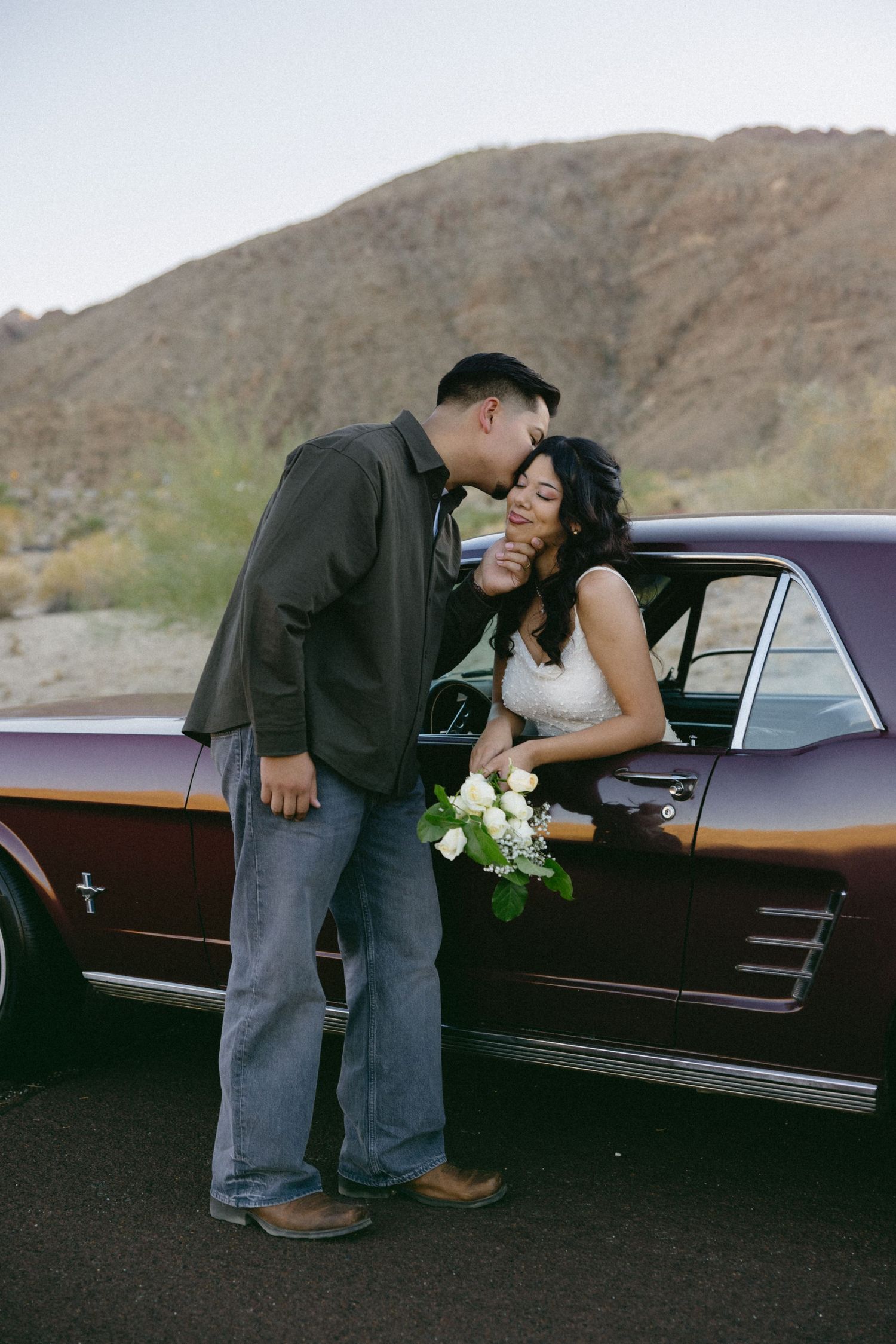 Romantic couple posing with vintage car in desert landscape with mountains in background.
